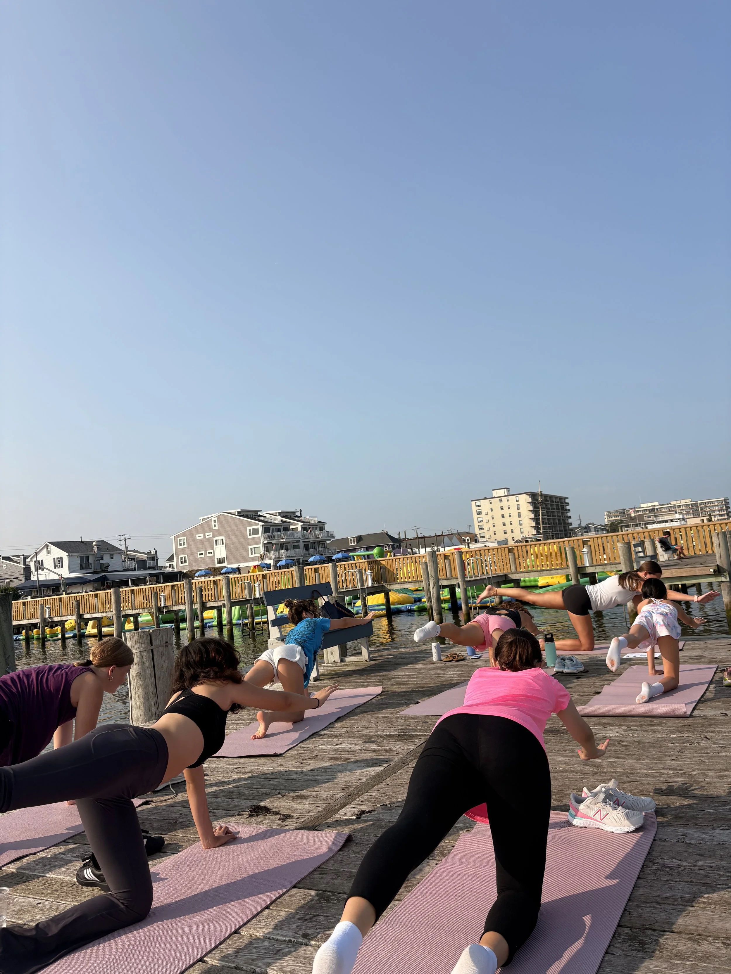 People practicing yoga on pink mats on a wooden pier by the water, with buildings and a clear sky in the background.