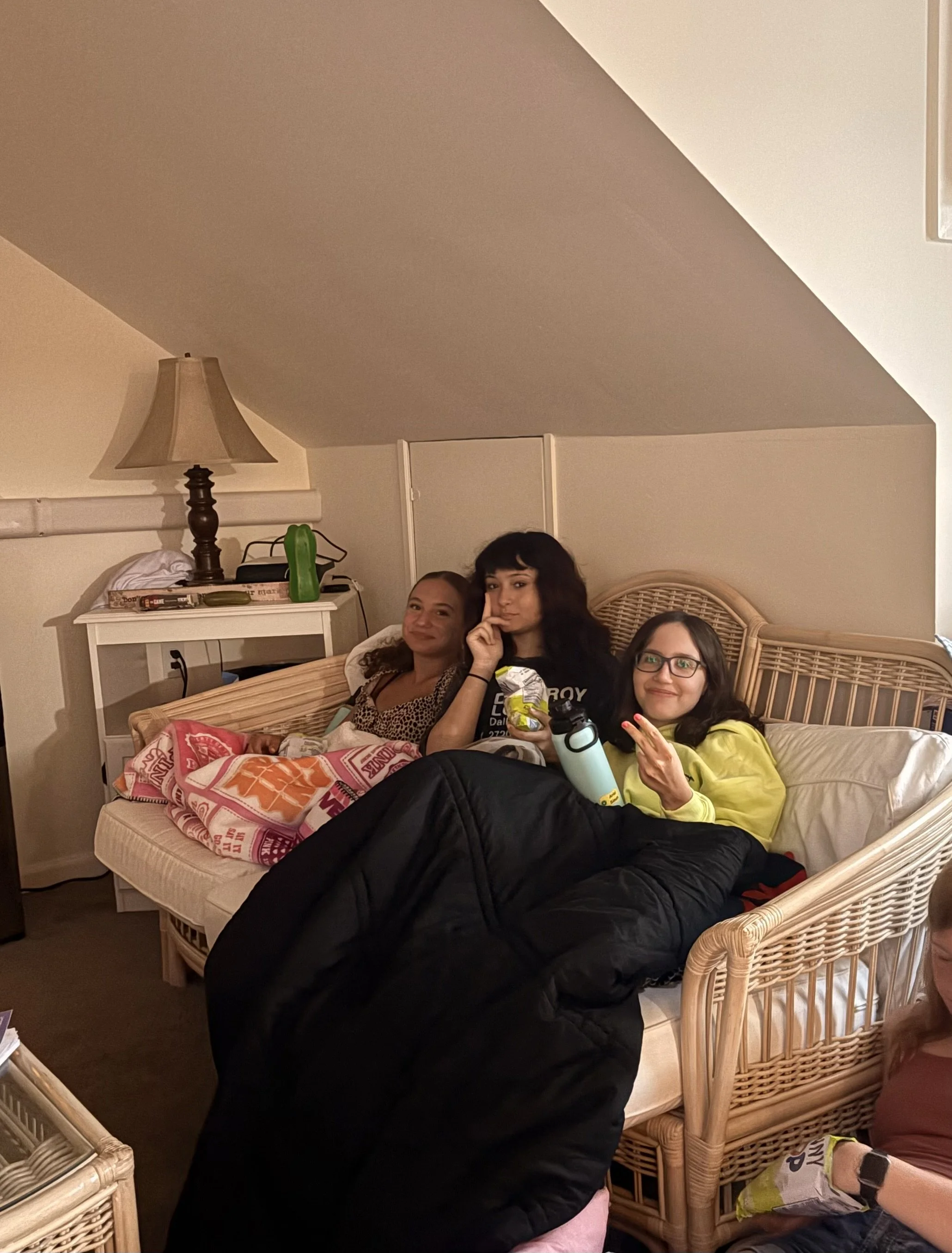 Four women sitting in a wicker bed, with one woman lying down and others sitting up, in a room with beige walls and furniture, some holding snacks and drinks, with a pillow and blanket on the bed.