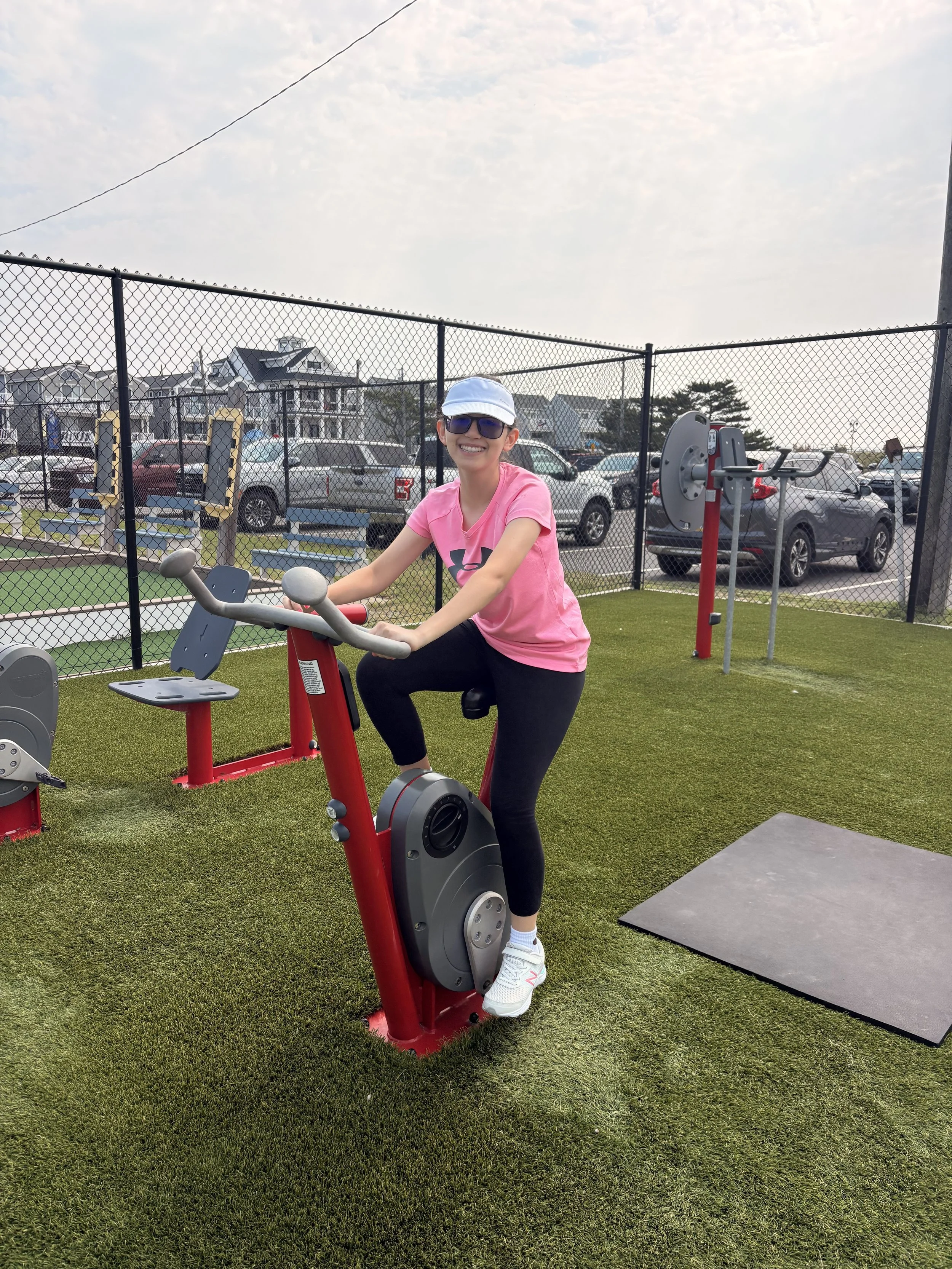 A smiling woman wearing a pink shirt, white cap, sunglasses, and white sneakers riding a red stationary exercise bike in an outdoor gym with green grass and a chain-link fence, parking lot, and houses in the background.