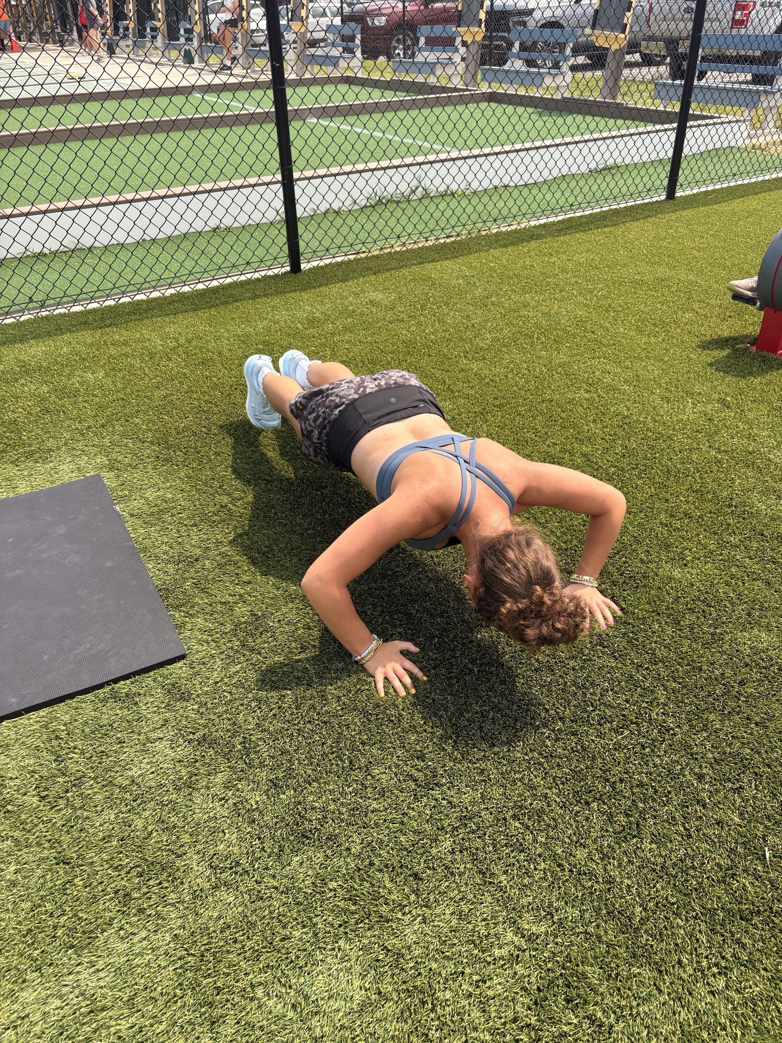 A woman doing a plank exercise on artificial grass outdoors at a park or sports facility with a chain-link fence and bleachers in the background.