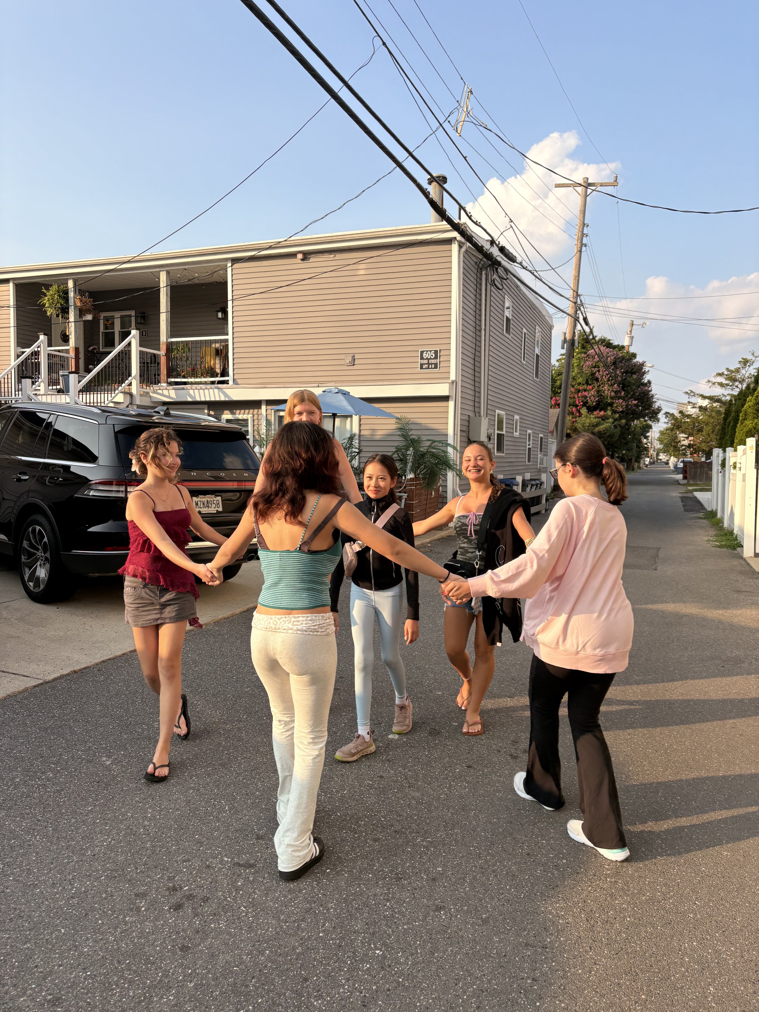 Group of young women holding hands, dancing or playing in a circle on a residential street during sunset, with a house, car, and utility poles in the background.
