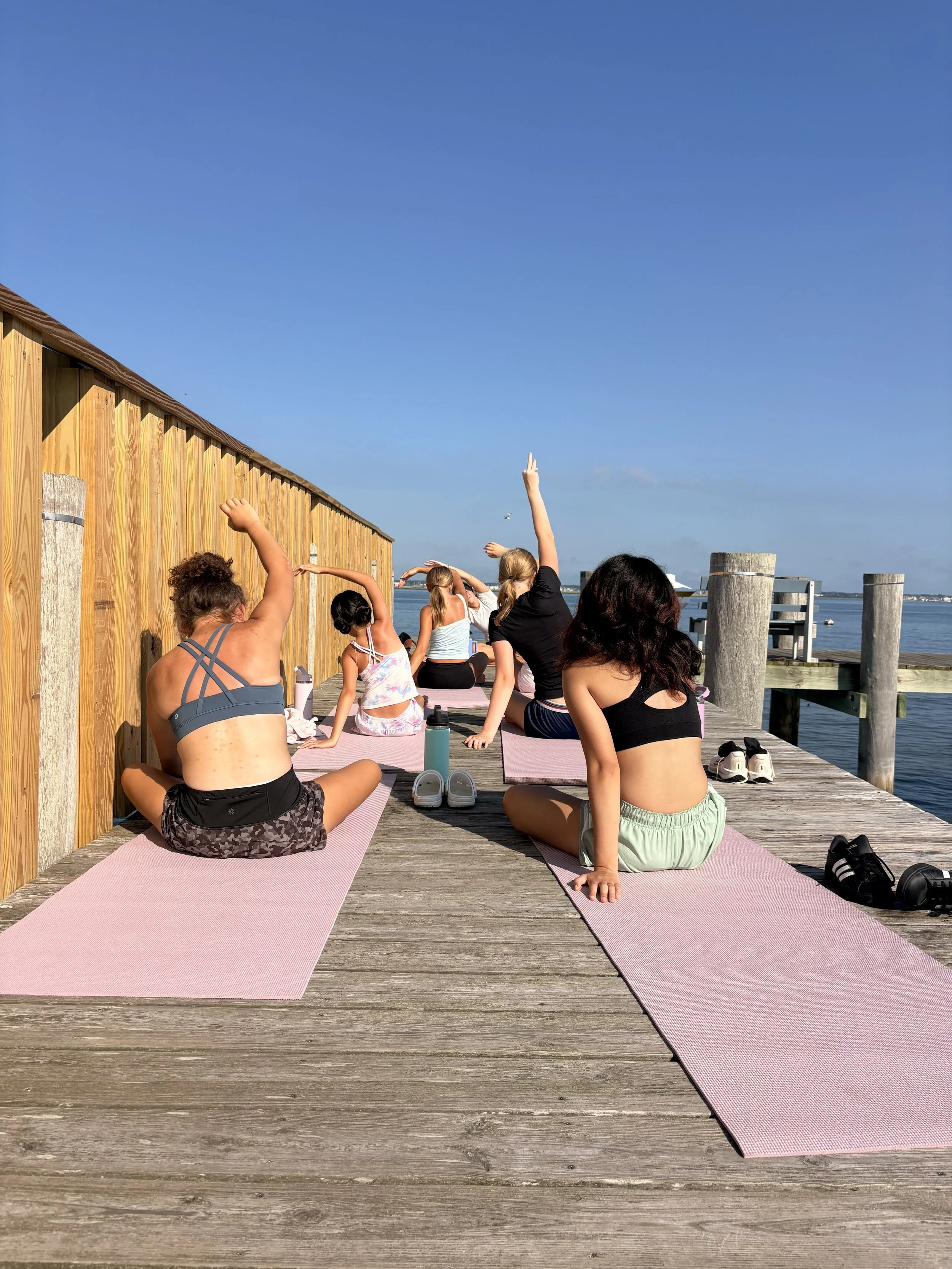 People participating in an outdoor yoga class on a wooden dock, practicing seated stretches and poses with water and a blue sky in the background.