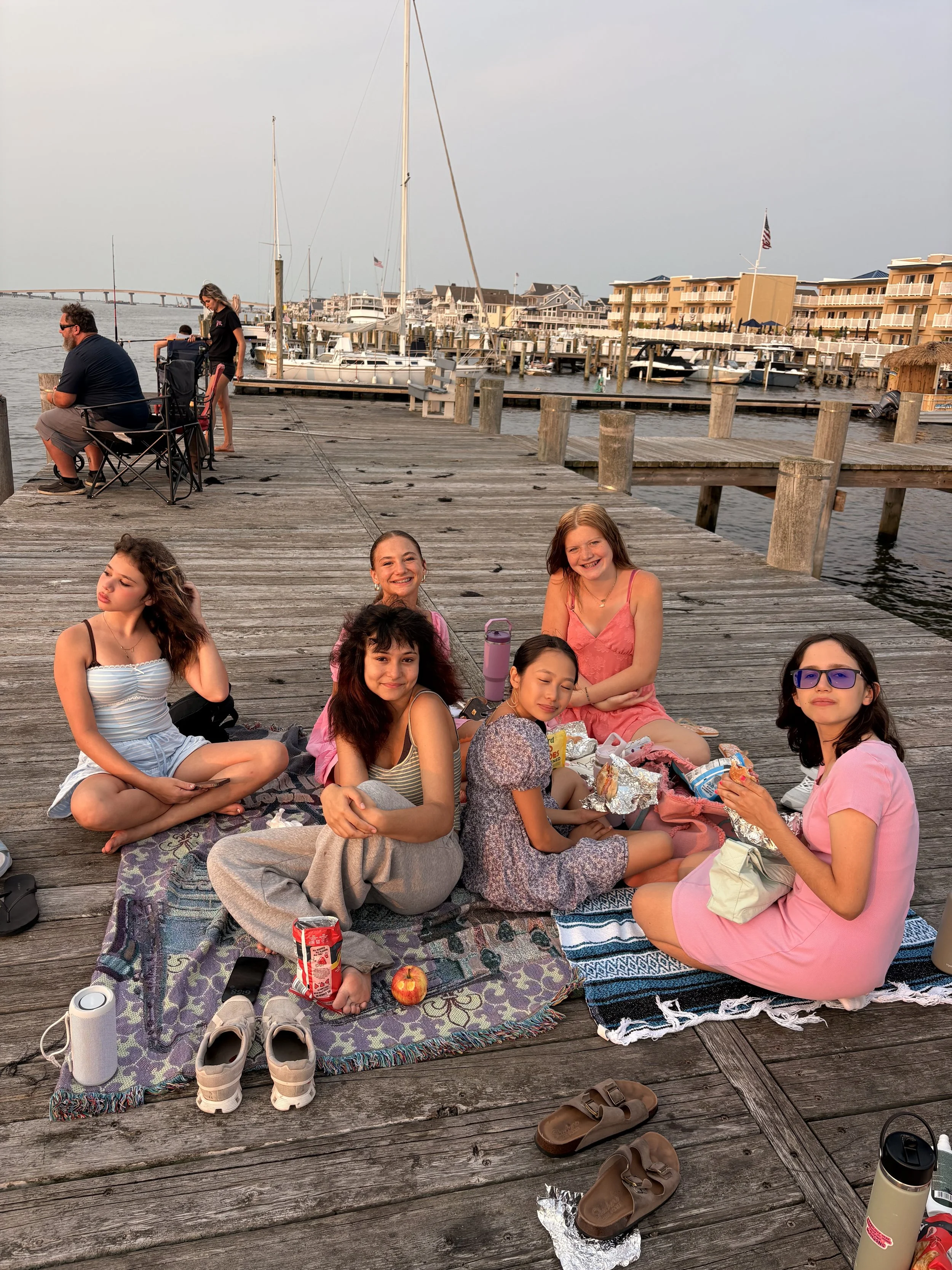 A group of children sitting on a blanket on a wooden dock by the water, enjoying a picnic with snacks and apple juices. In the background, there are boats and houses along the waterfront.