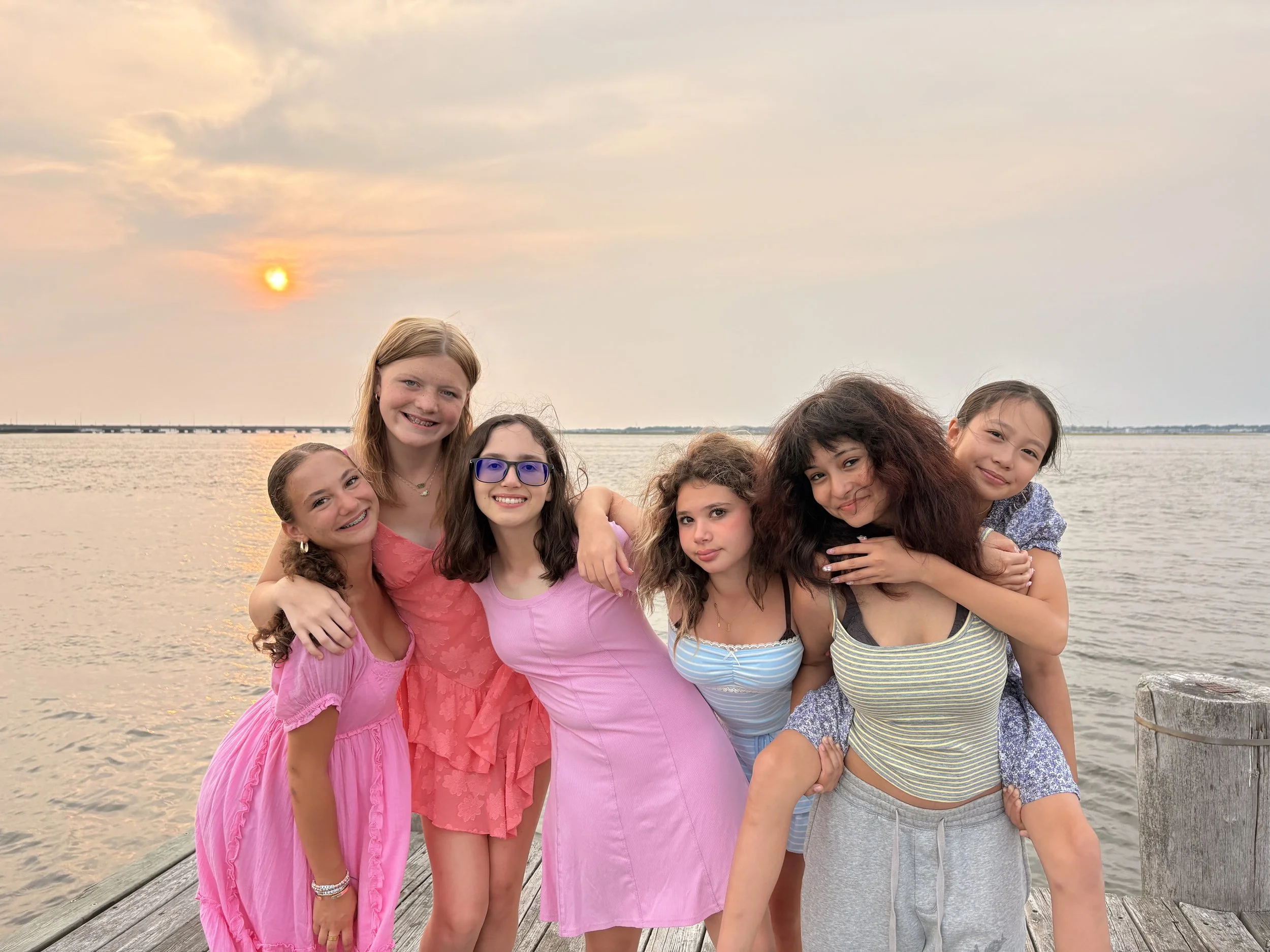 A group of eight girls smiling and hugging each other on a wooden dock by the water during sunset