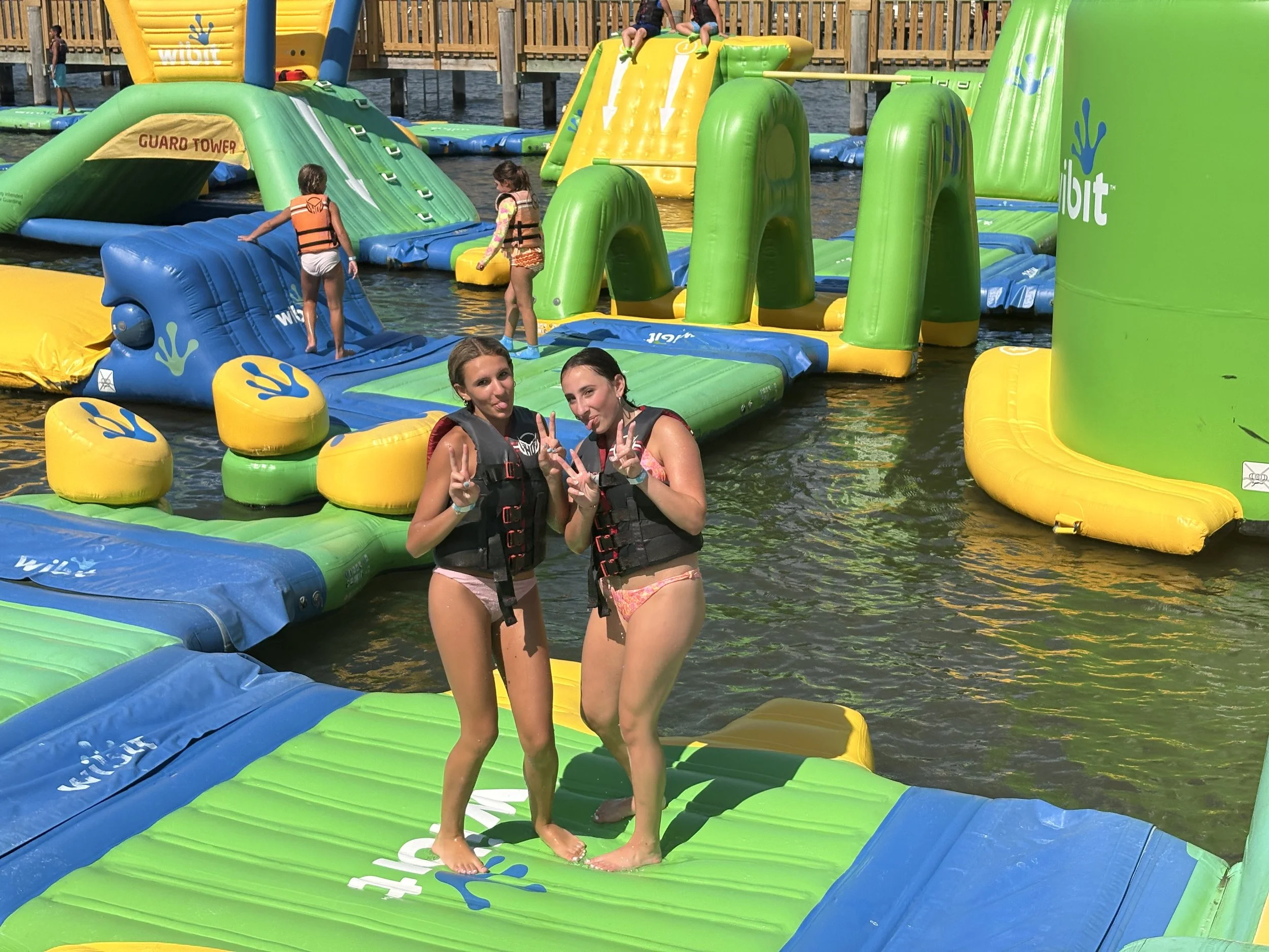 Two young girls in life jackets standing on an inflatable water obstacle course, making peace signs and sticking out their tongues, while other children play on the course in the background near a wooden dock.