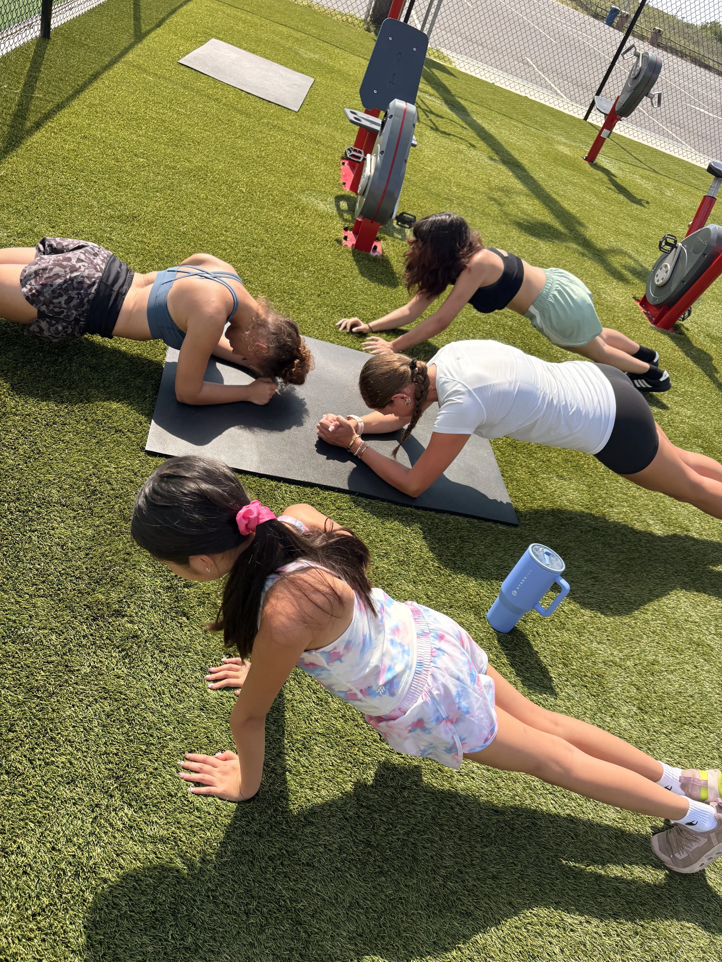 A group of young girls doing push-ups on a grassy field at an outdoor sports facility, with sports equipment and fenced courts in the background.