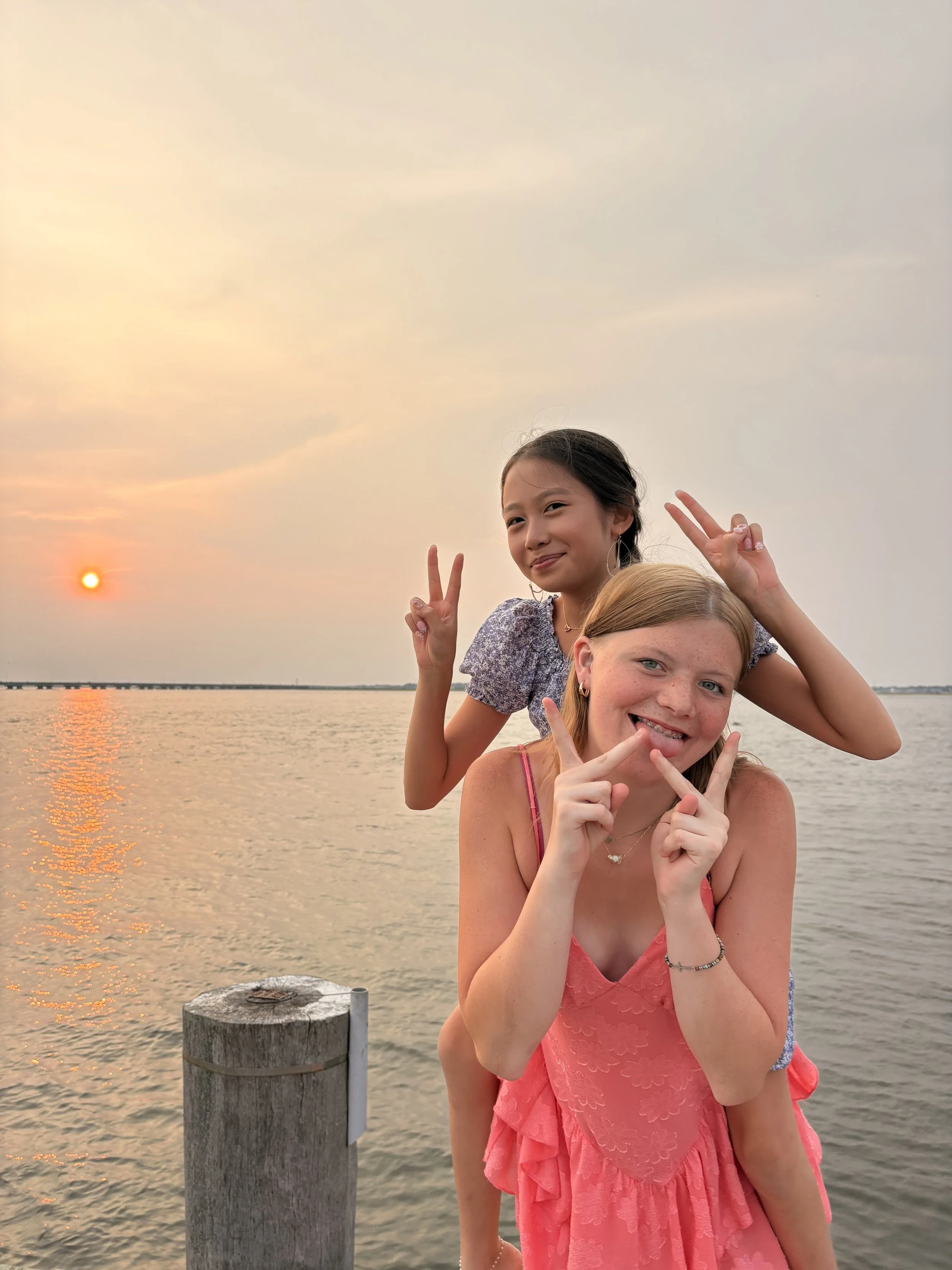 Two girls smiling and posing with peace signs near a body of water at sunset, with a cloudy sky and a dock in the background.