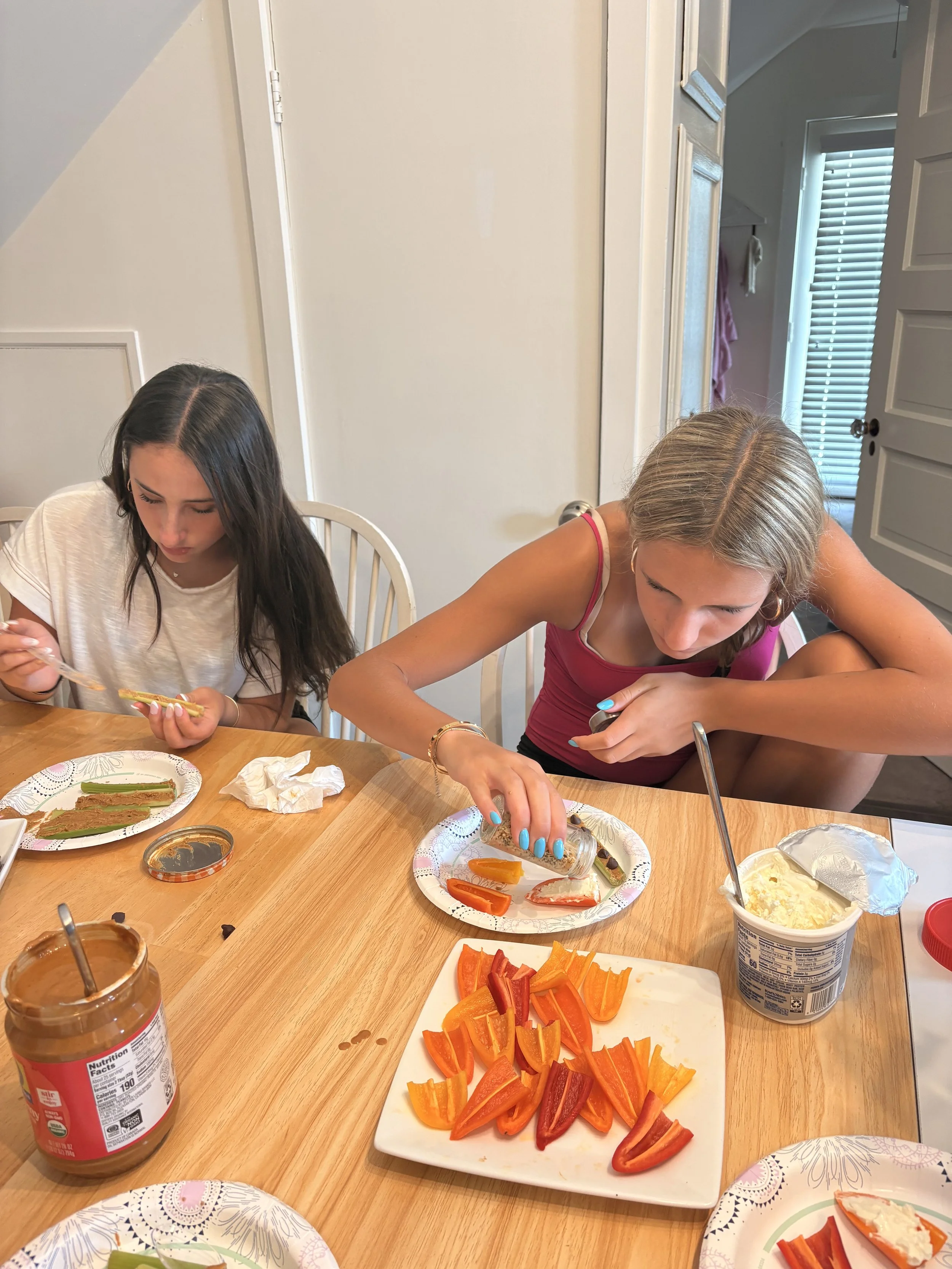 Two girls sitting at a wooden table with food and condiments, one pouring dressing on a plate of sliced colorful bell peppers, the other looking down at her plate, in a bright room with a white door and window with blinds in the background.