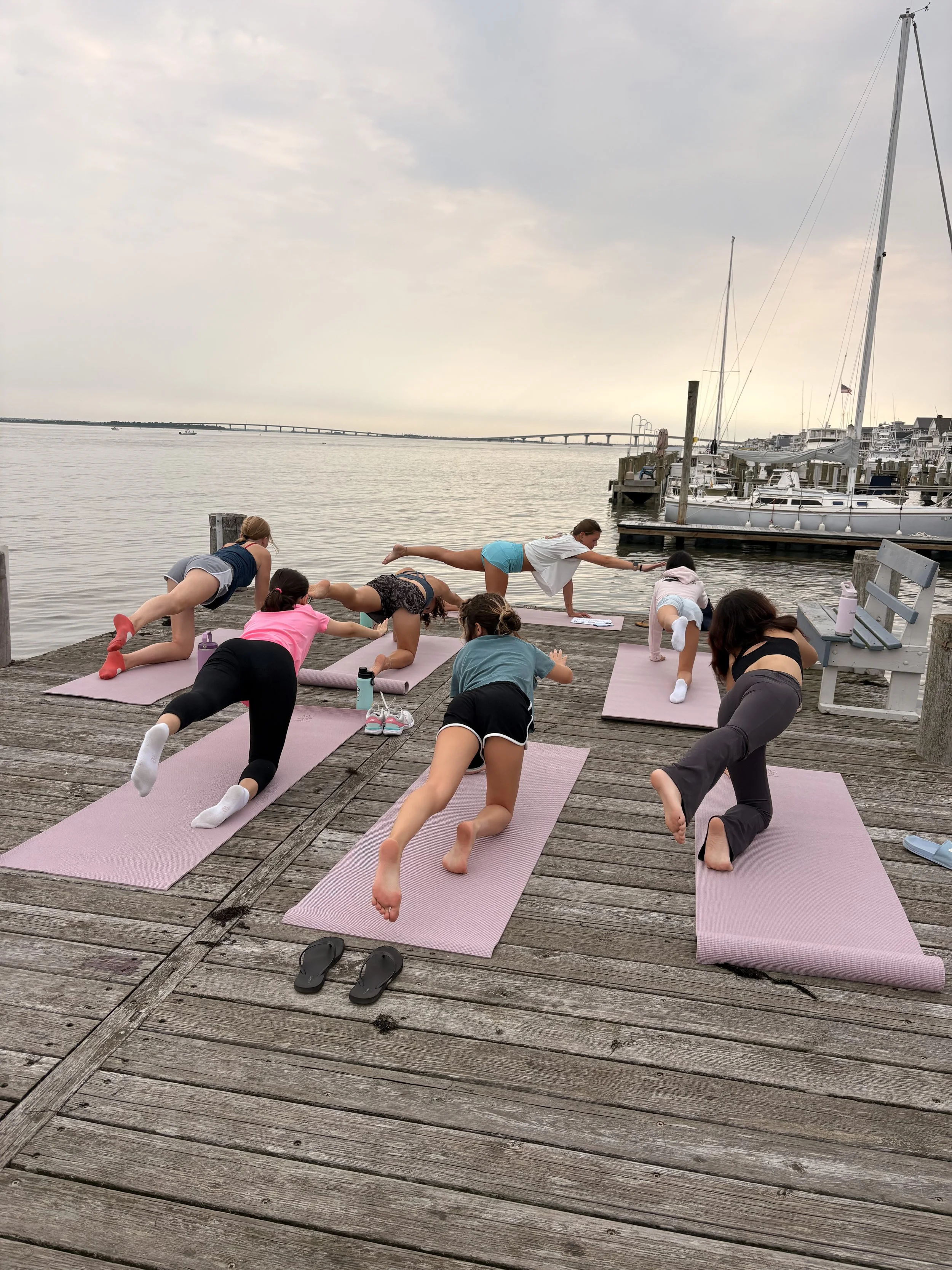 People practicing yoga outdoors on a wooden dock by the water, with sailboats and a bridge in the background on a cloudy day.