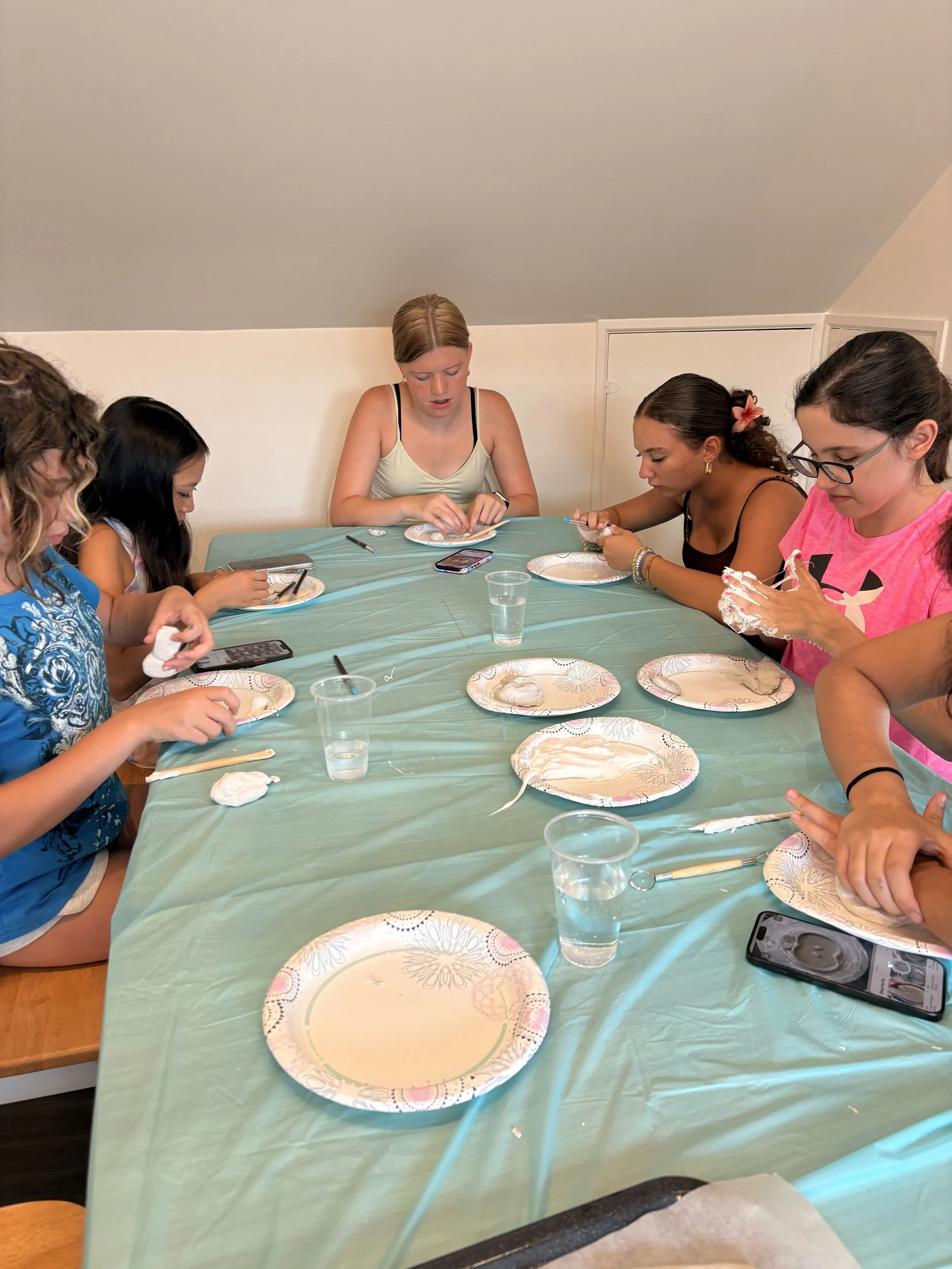 Young women sitting at a table covered with a green tablecloth, engaging in decorating activity with paper plates, using tools and their hands, some with smartphones nearby.