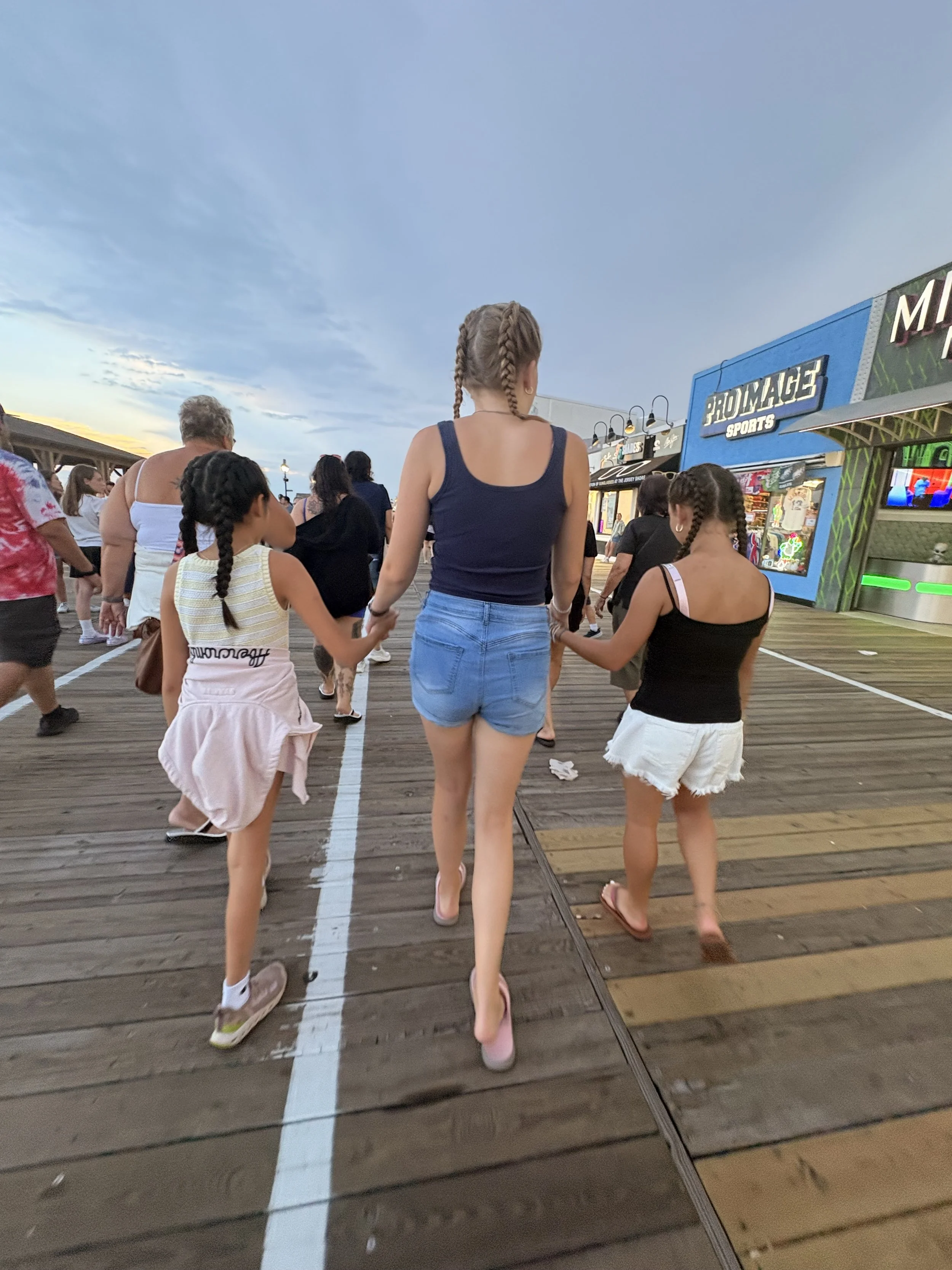 People walking on a wooden boardwalk at sunset, with shops and game booths in the background and a cloudy sky above.