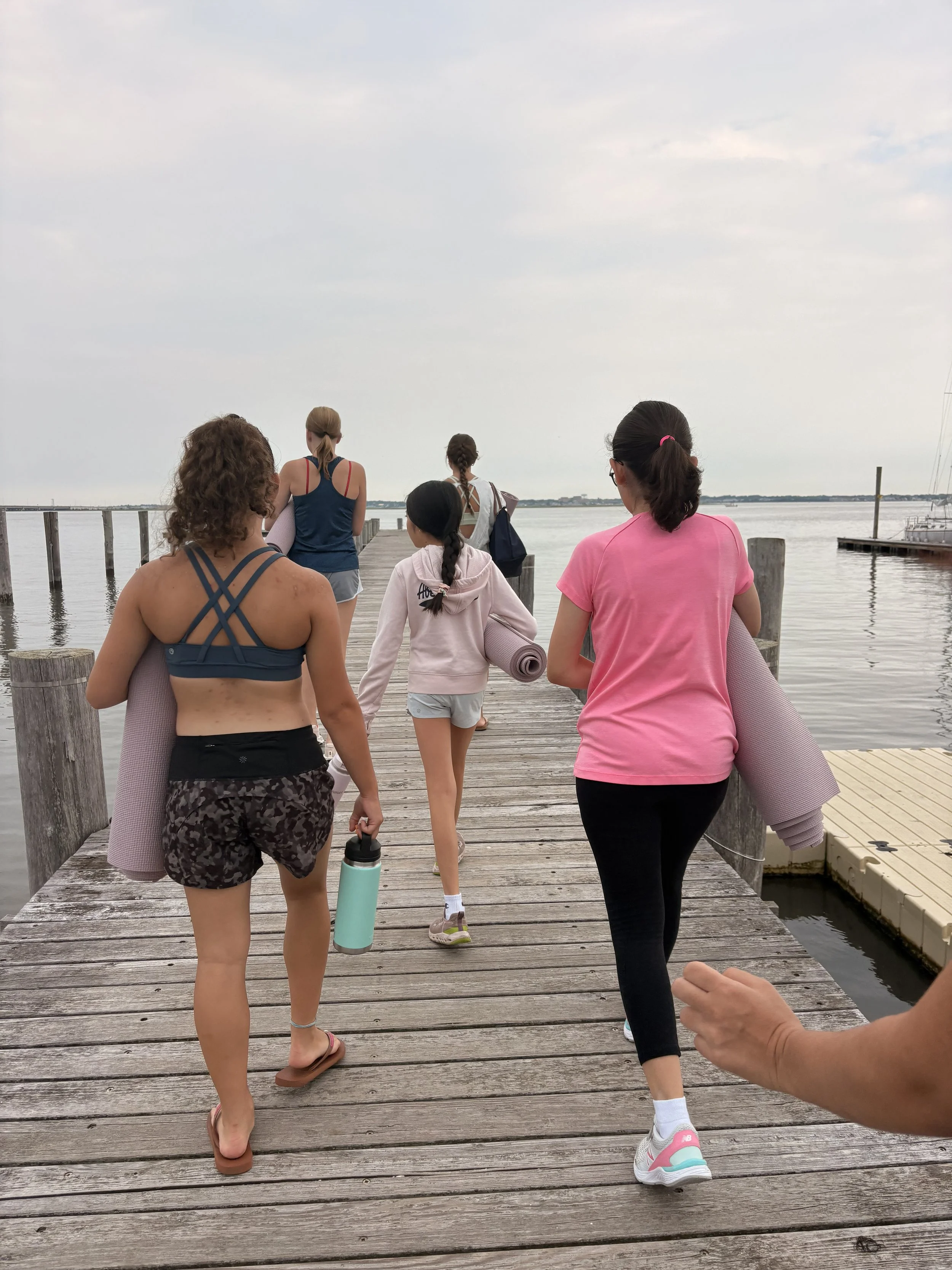 Group of women walking on a wooden dock by the water, some carrying yoga mats, during overcast weather.