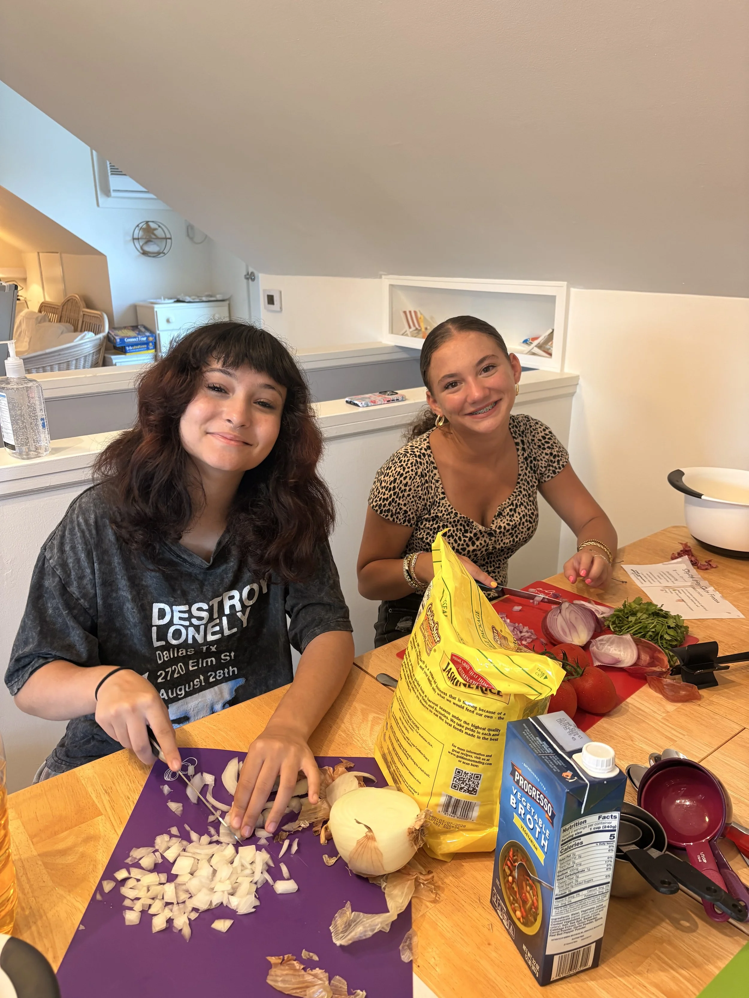 Two young women sitting at a dining table preparing vegetables for cooking. One woman with shoulder-length dark hair wearing a black t-shirt, and the other with long brown hair and braces wearing a leopard print top. The table has chopped onions, a w