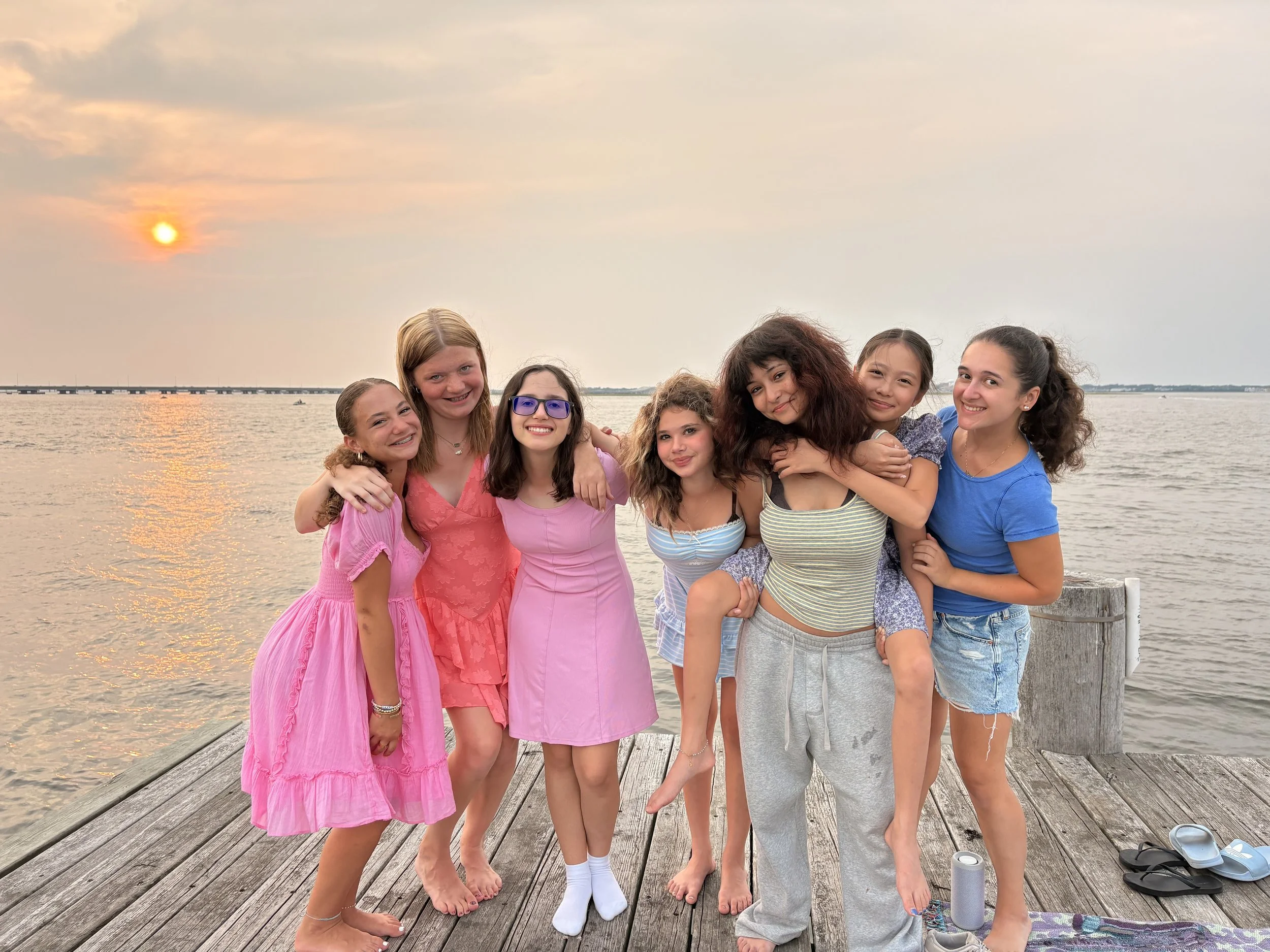Group of seven girls standing arm-in-arm on a wooden dock by the water during sunset, smiling and wearing casual summer clothing.