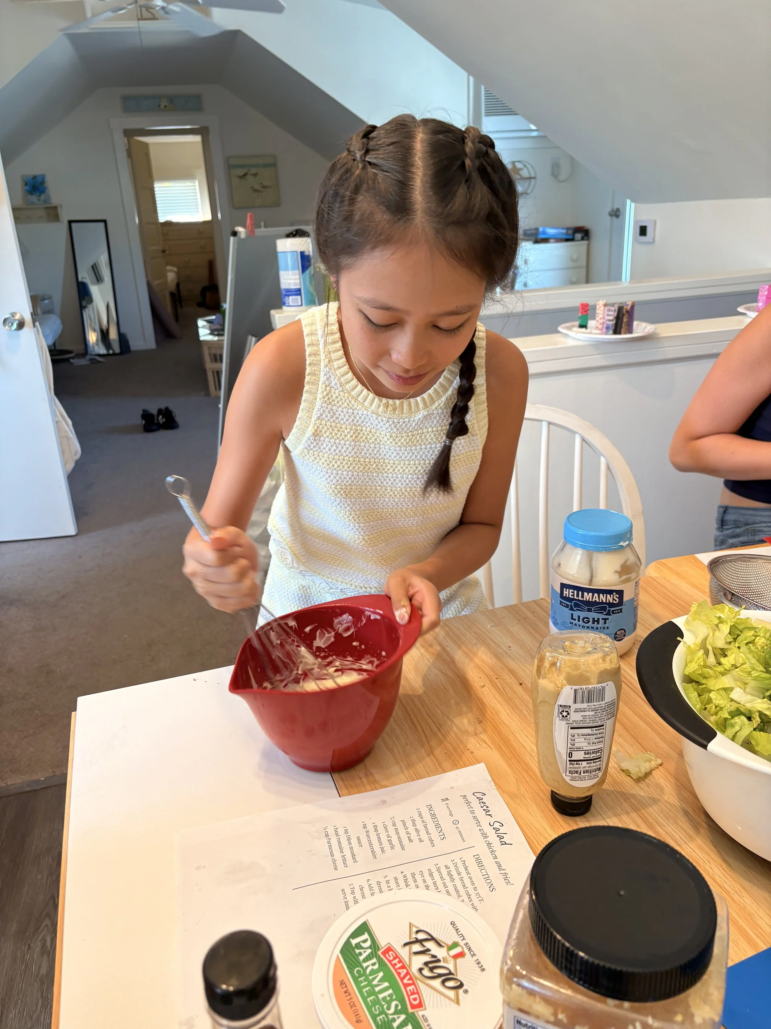 A young girl with braided hair is sitting at a table, preparing to make a salad with a red bowl, lettuce, and a jar of mayonnaise. Other salad ingredients and condiments are also on the table.