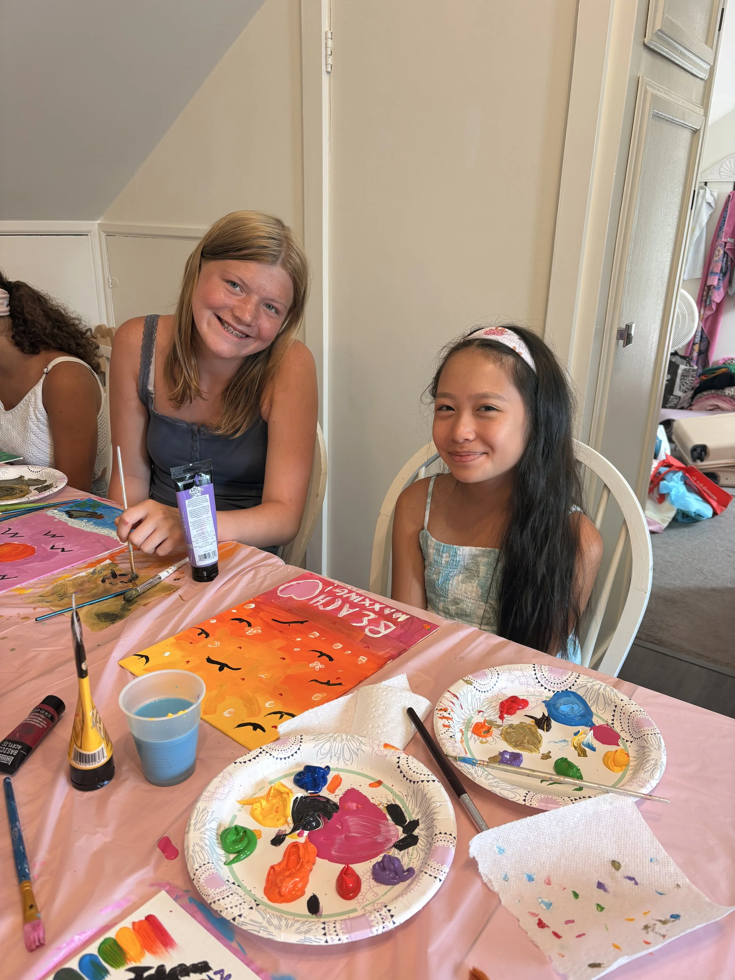 Two young girls sitting at a table with painting supplies, including paint palettes, brushes, and a painting with Halloween-themed colors and bats. The girl on the left has red hair and braces, smiling, while the girl on the right has dark hair with 