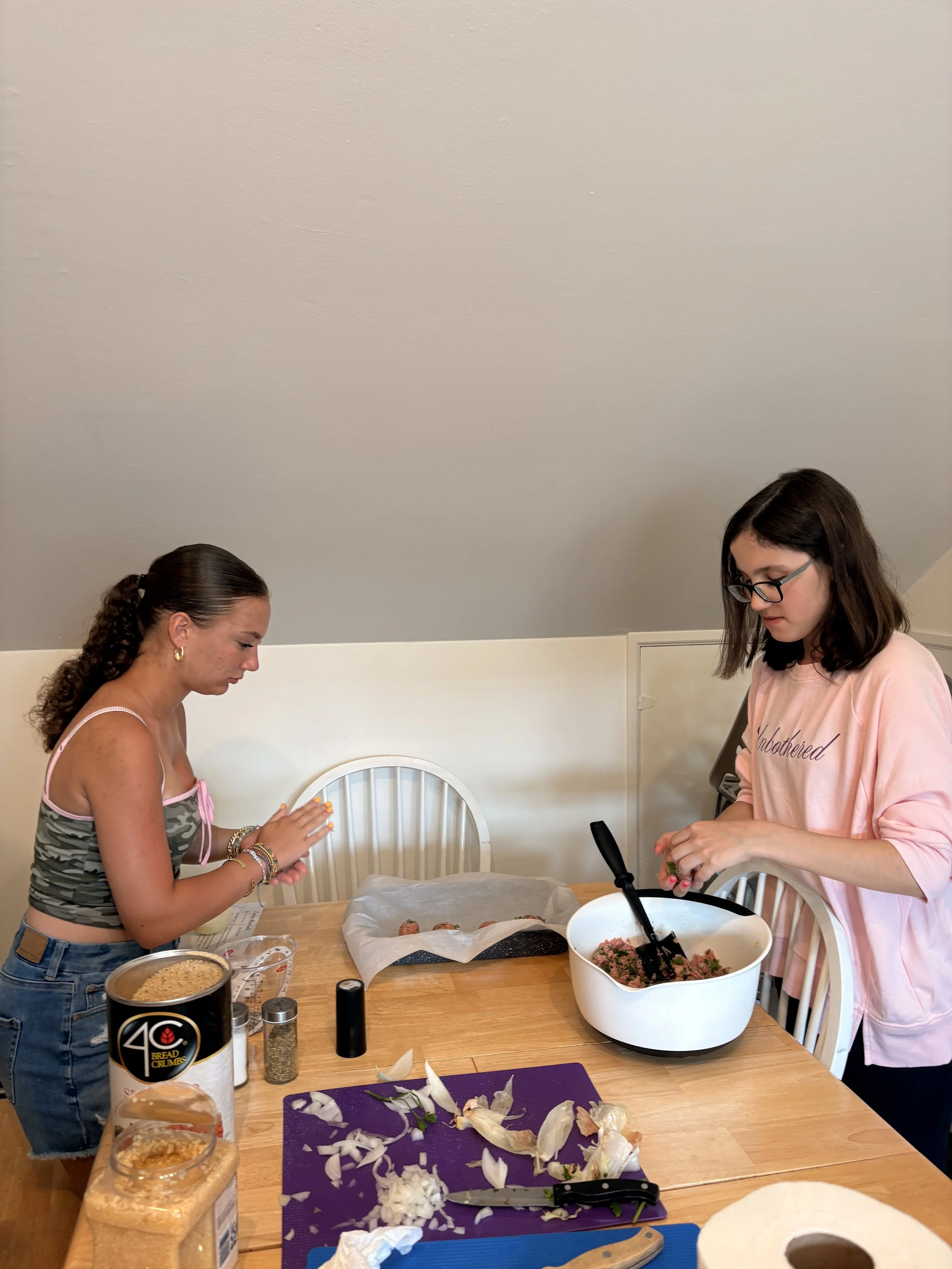 Two women are preparing food in a kitchen at a wooden dining table with ingredients and cooking utensils. One woman has curly brown hair and is wearing a sleeveless top, while the other has straight dark hair, glasses, and is wearing a pink shirt.