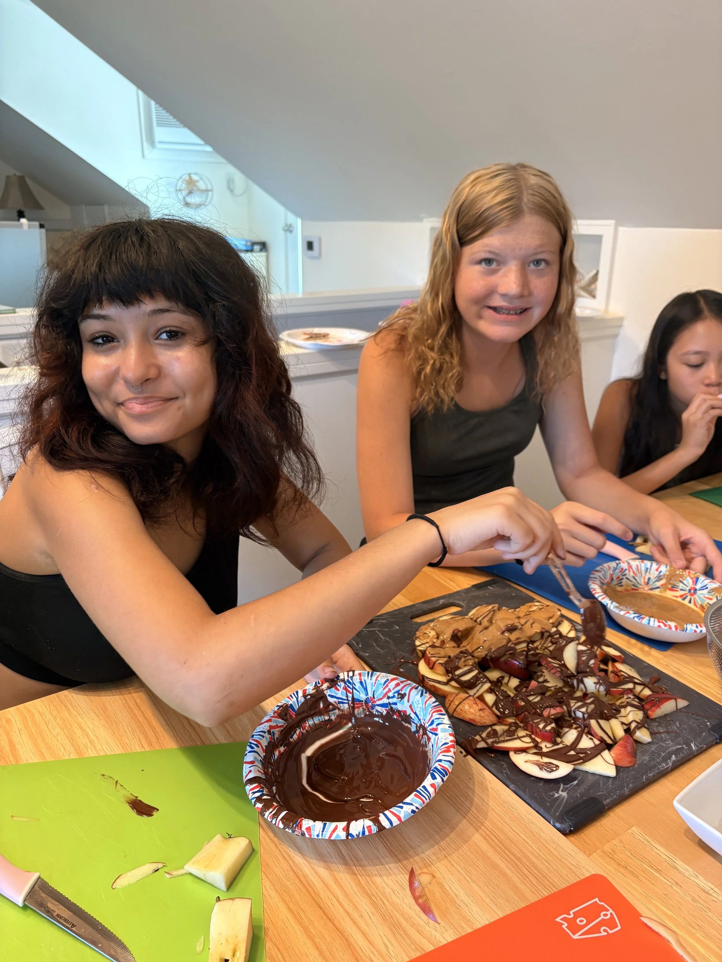 Three young women, with one in the foreground smiling, preparing and decorating a large tray of sliced fruit and cookies with chocolate sauce at a kitchen table.