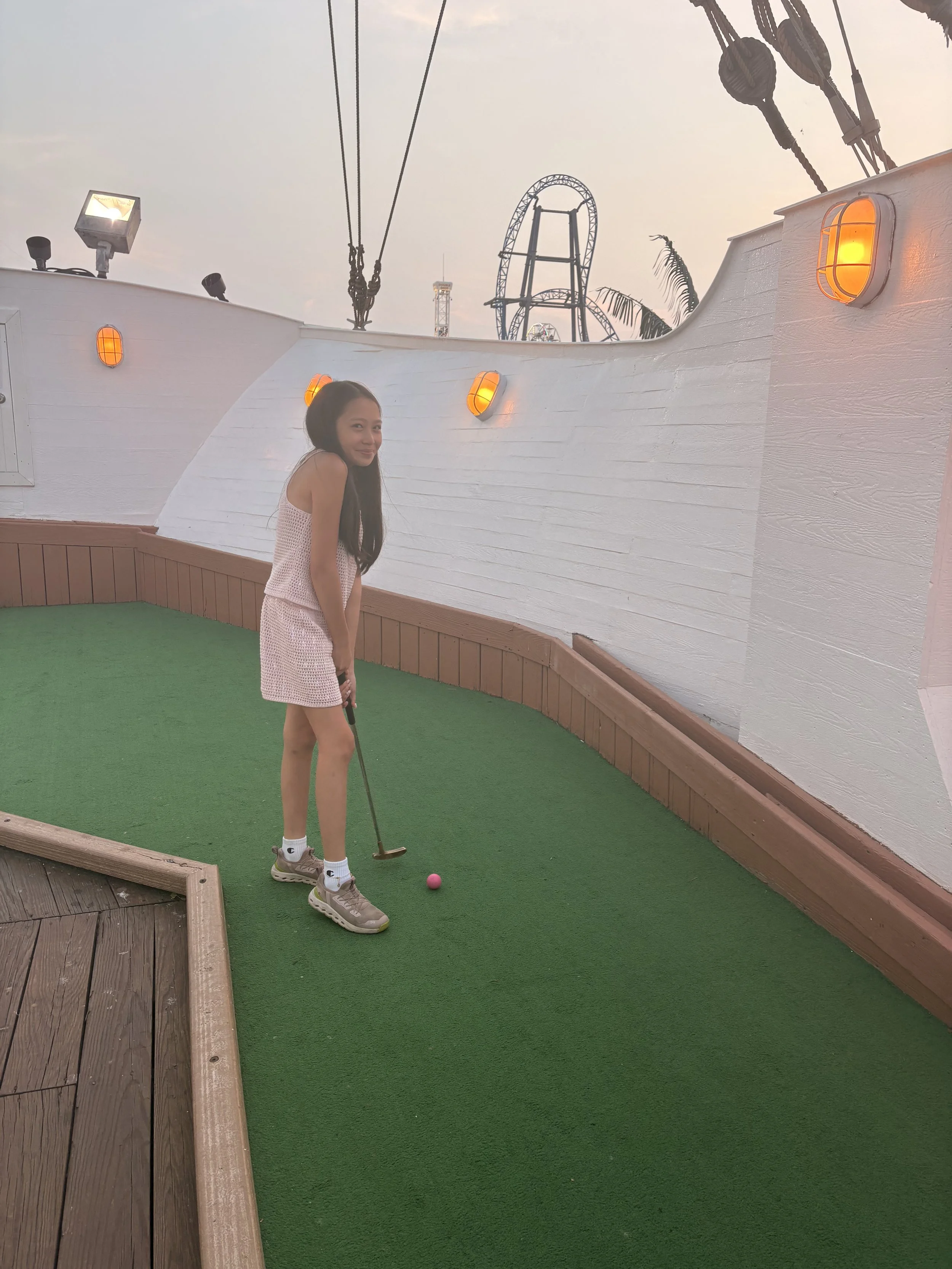 A young girl with long dark hair stands on a miniature golf course, preparing to hit a pink golf ball with a putter. She is wearing a pink dress, white socks, and beige sneakers. The background shows amusement park rides, including a roller coaster, 