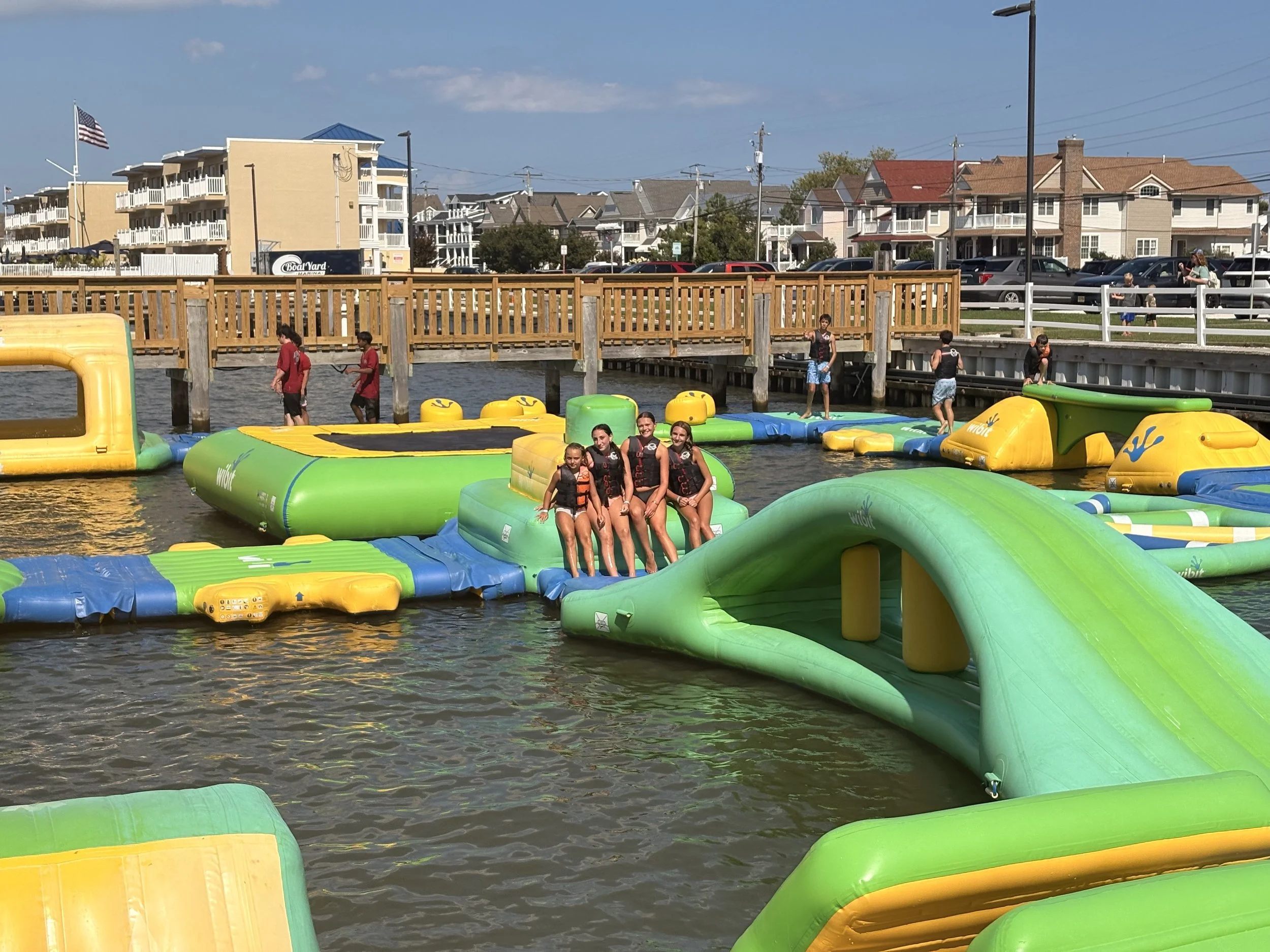 People enjoying on water inflatables at a lake, with a wooden pier, houses, and parked cars in the background.