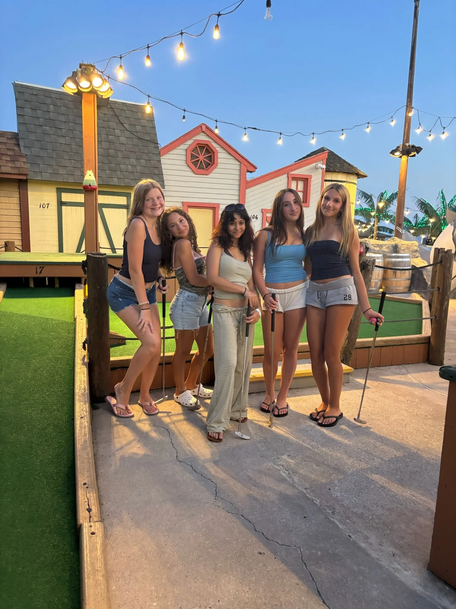 A group of five young women posing with mini golf clubs on a miniature golf course. They are standing in front of small colorful house structures, with string lights hanging overhead and palm trees in the background during the evening.