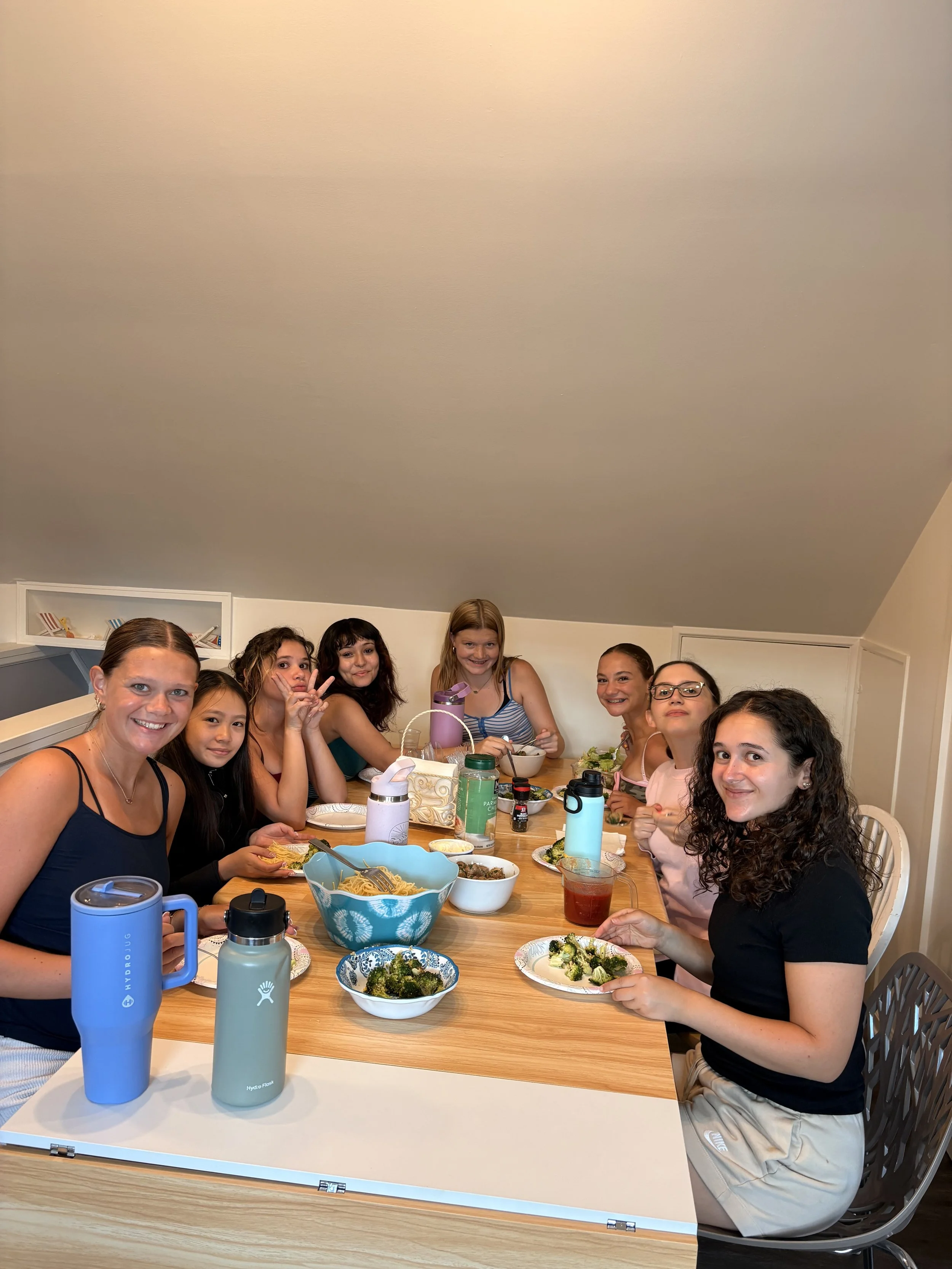 A group of young girls and women sitting around a dining table enjoying a meal, with bowls of salad and noodles in front of them.
