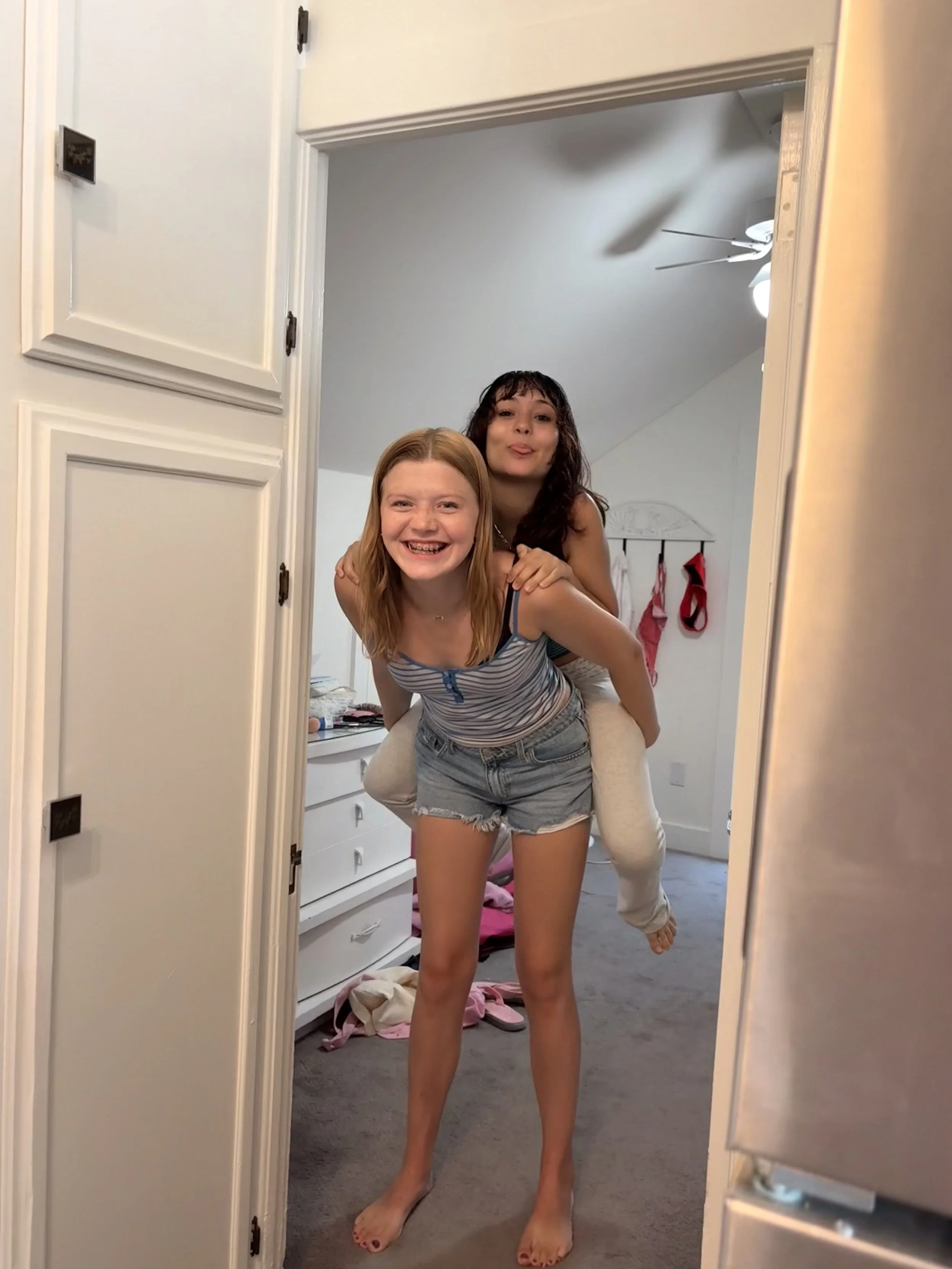 Two young women with brown and red hair, smiling, playfully posing in front of a mirror in a bedroom. One is giving a piggyback ride to the other. The room has a white dresser and clothes on the floor.