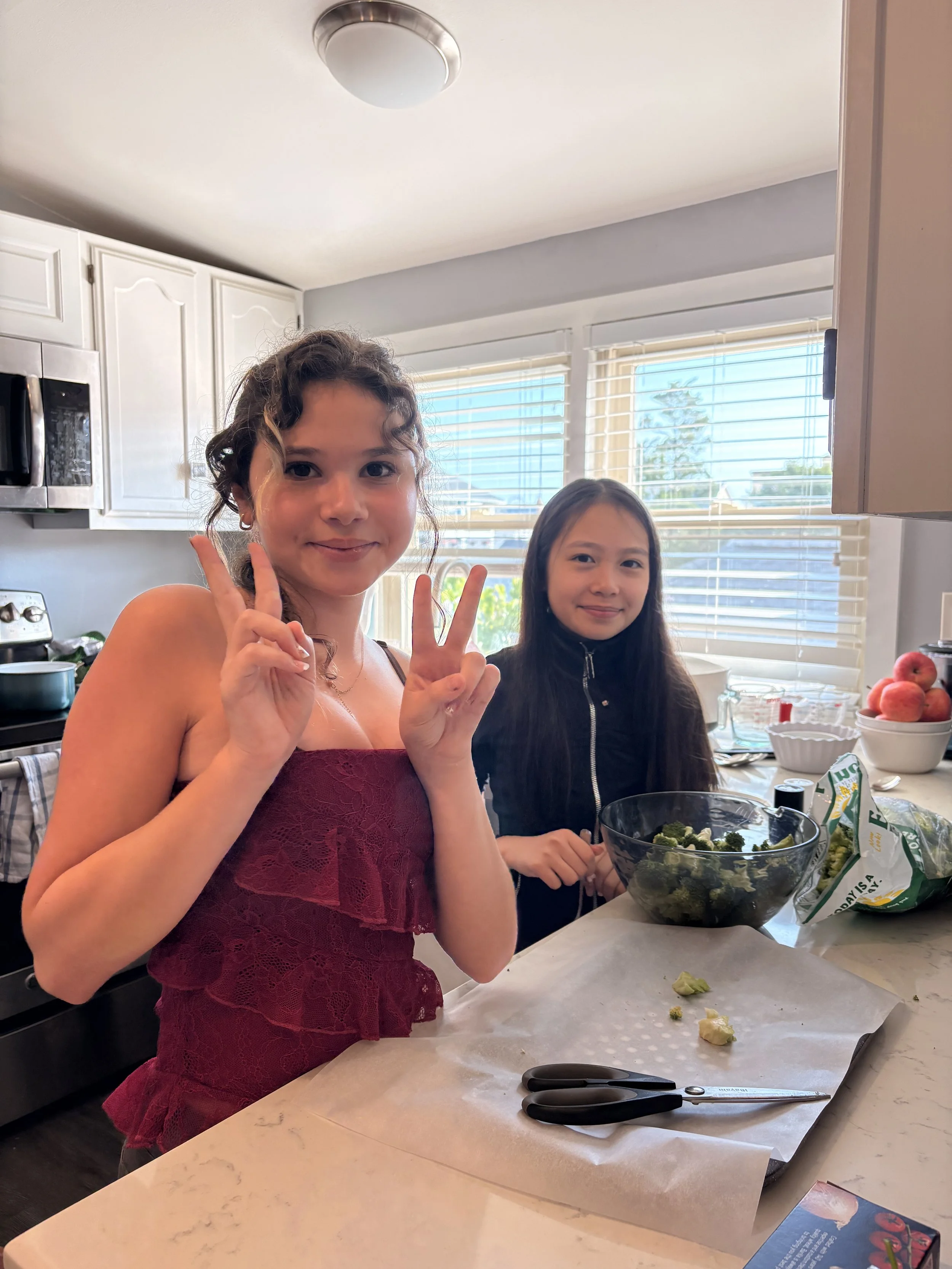 Two girls making a peace sign pose in a kitchen with a bowl of broccoli on the counter