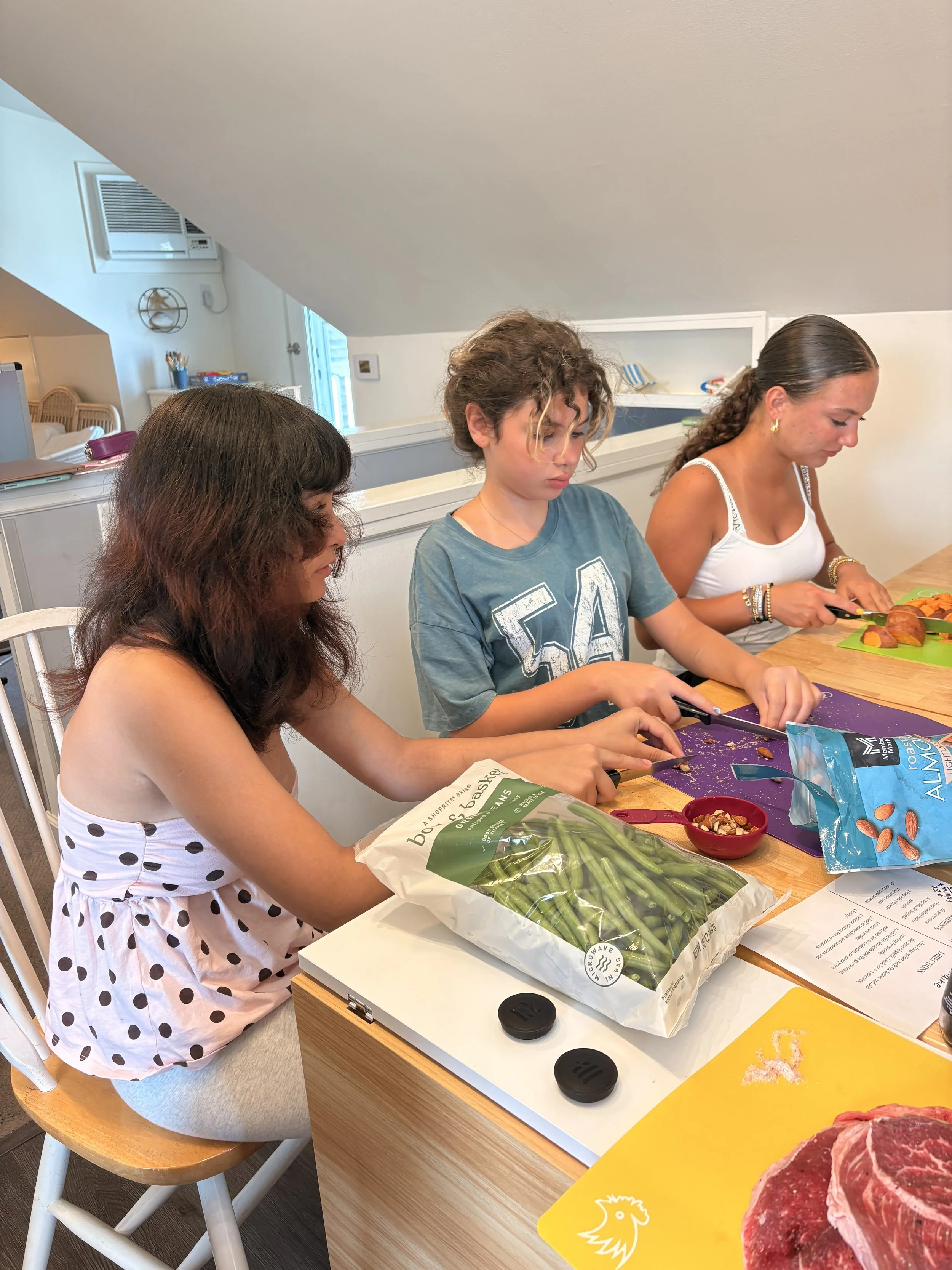 Three women are preparing food at a kitchen table with various ingredients including green beans, sliced sweet potatoes, and raw meat, in a well-lit room with white walls and a sloped ceiling.