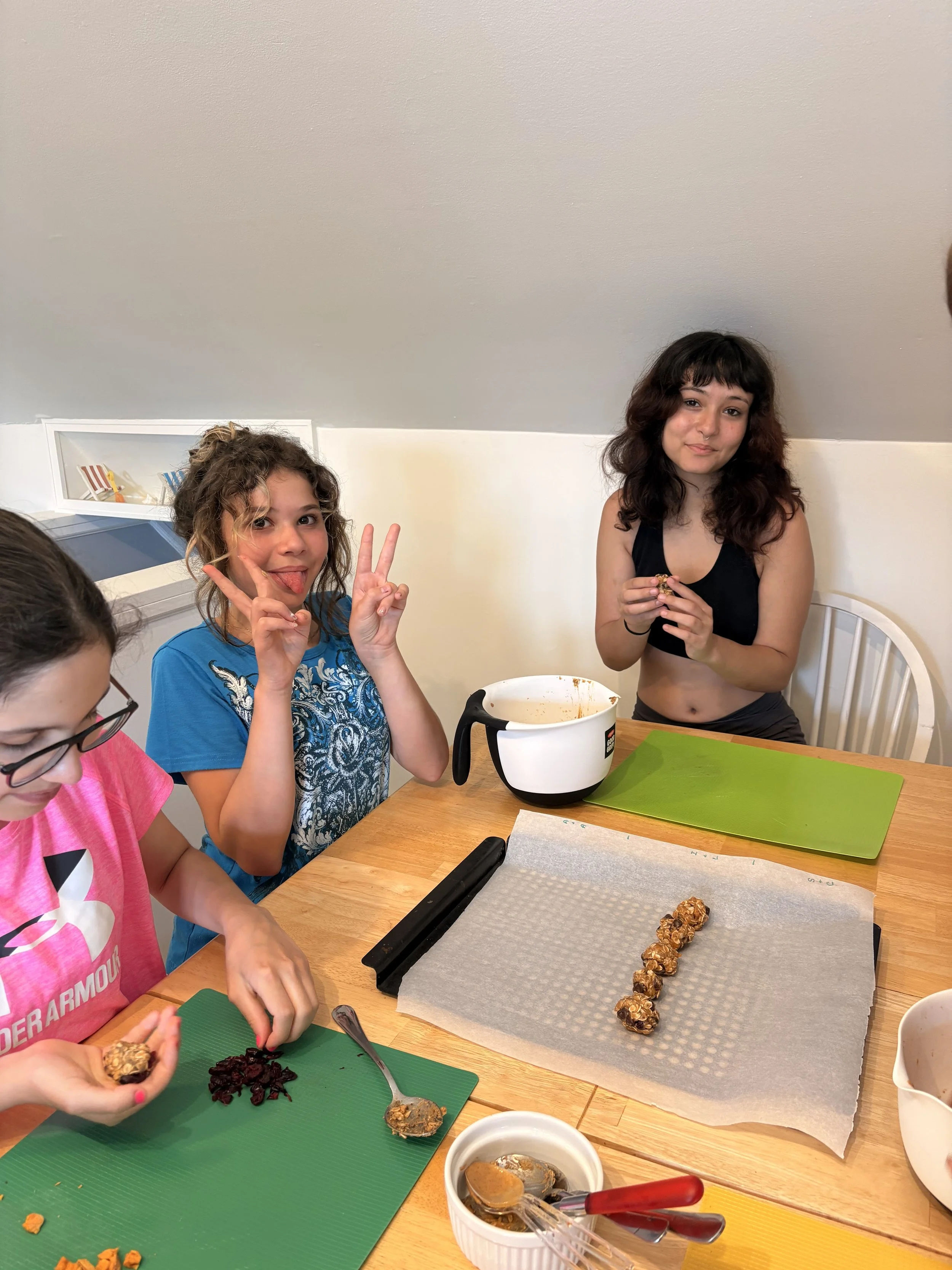 Three girls at a kitchen table preparing and eating homemade energy bites or granola bars. One girl holds a ball of mixture, another girl makes a peace sign, and the third girl looks at the camera. The table has green and yellow cutting mats, a bakin