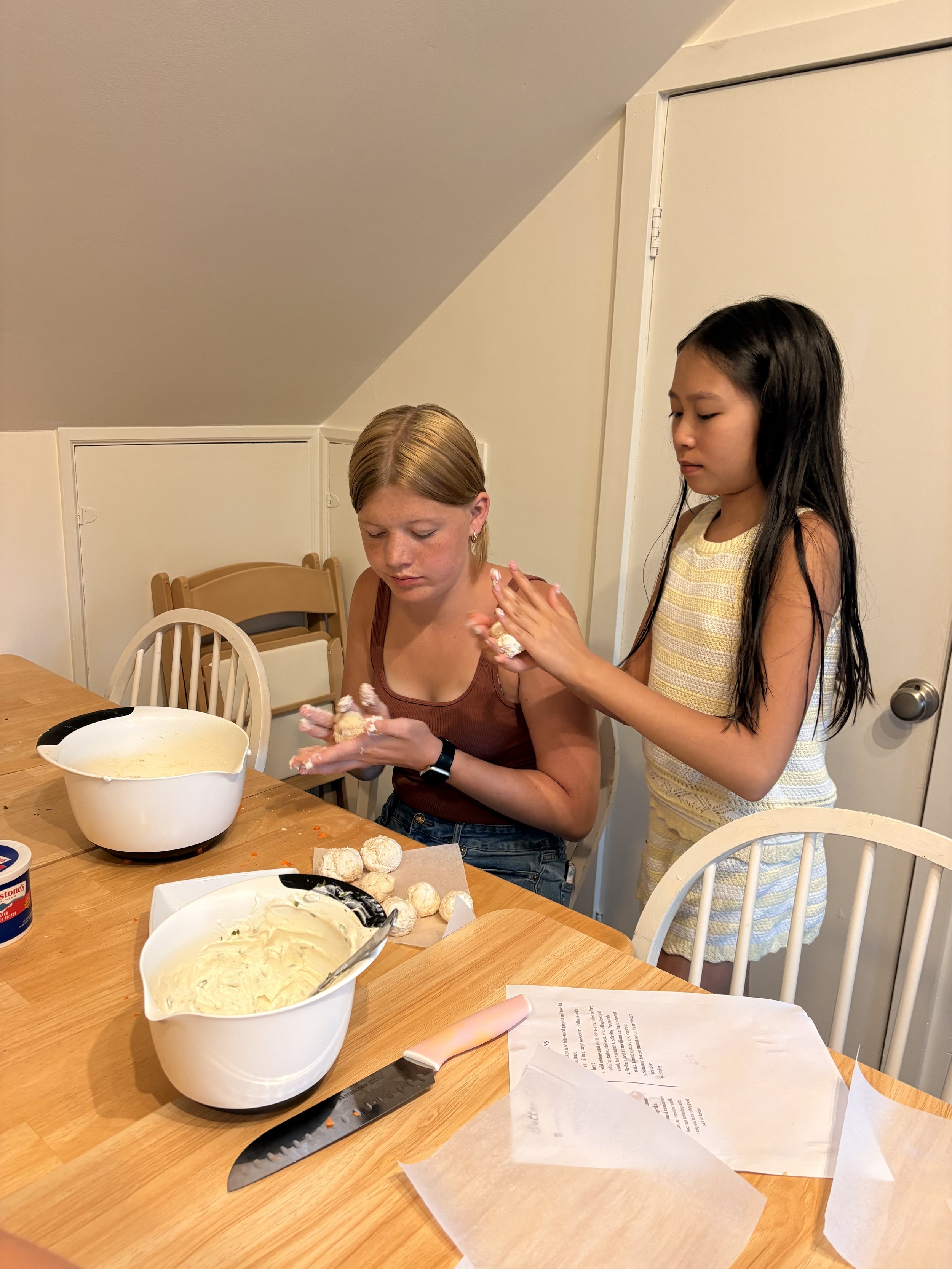 A young woman with red hair and a tank top sitting at a dining table, holding a dough ball. A young girl with long black hair and a sleeveless yellow dress is standing next to her, helping or giving her something. The table has bowls with dough and a