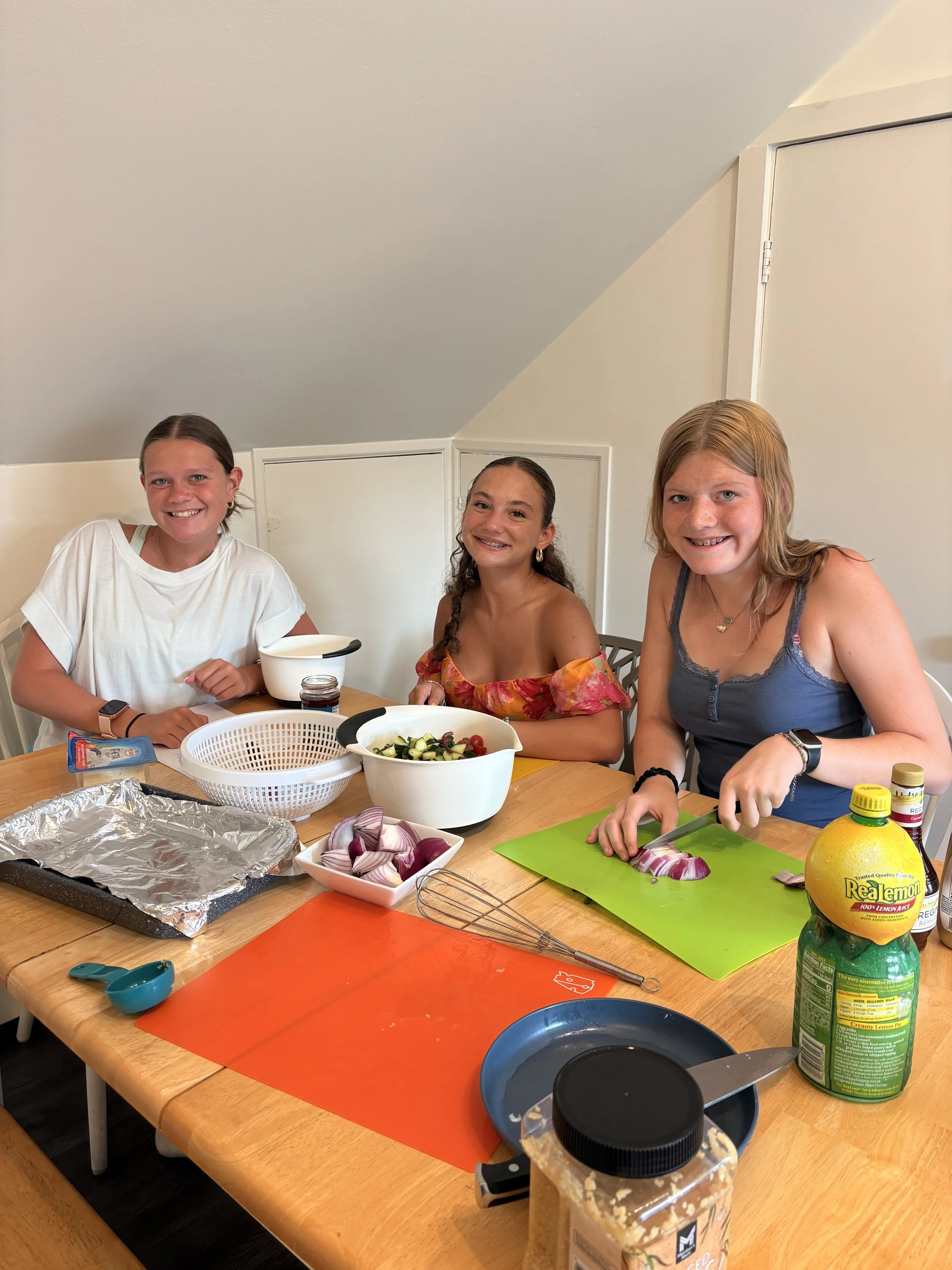 Three girls sitting at a kitchen table preparing food with vegetables, cutting onions, and other ingredients, surrounded by kitchen utensils and supplies.