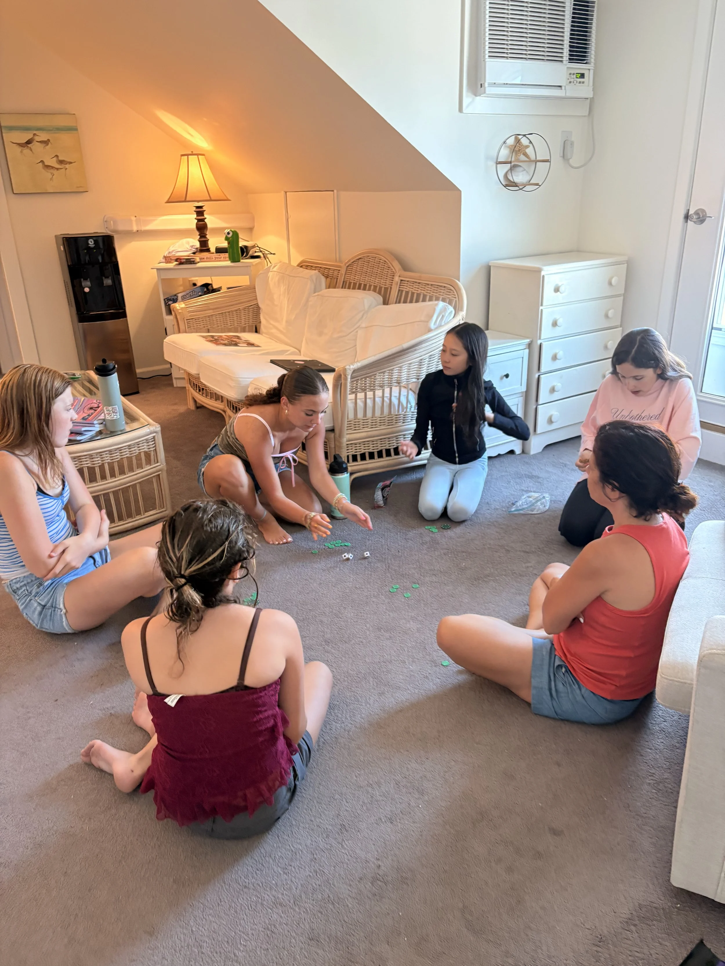 A group of seven people, mostly young females, sitting on the carpeted floor in a circle, playing a game with small green and white objects, in a cozy living room with a wicker sofa, a small white dresser, and various decorative items.