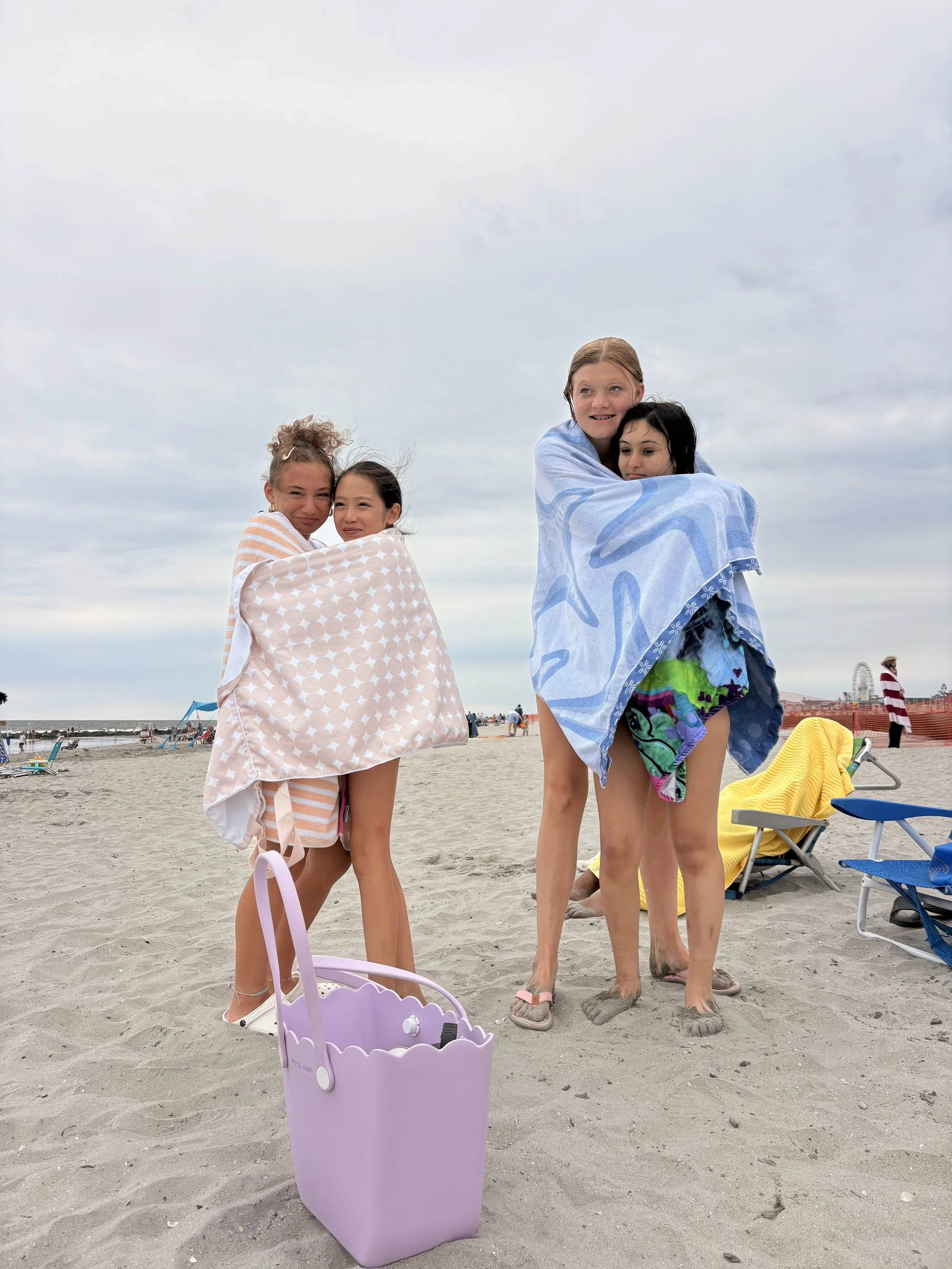 Four children wrapped in towels standing on a sandy beach, smiling and hugging each other, with a purple beach bag in the foreground and a cloudy sky overhead.
