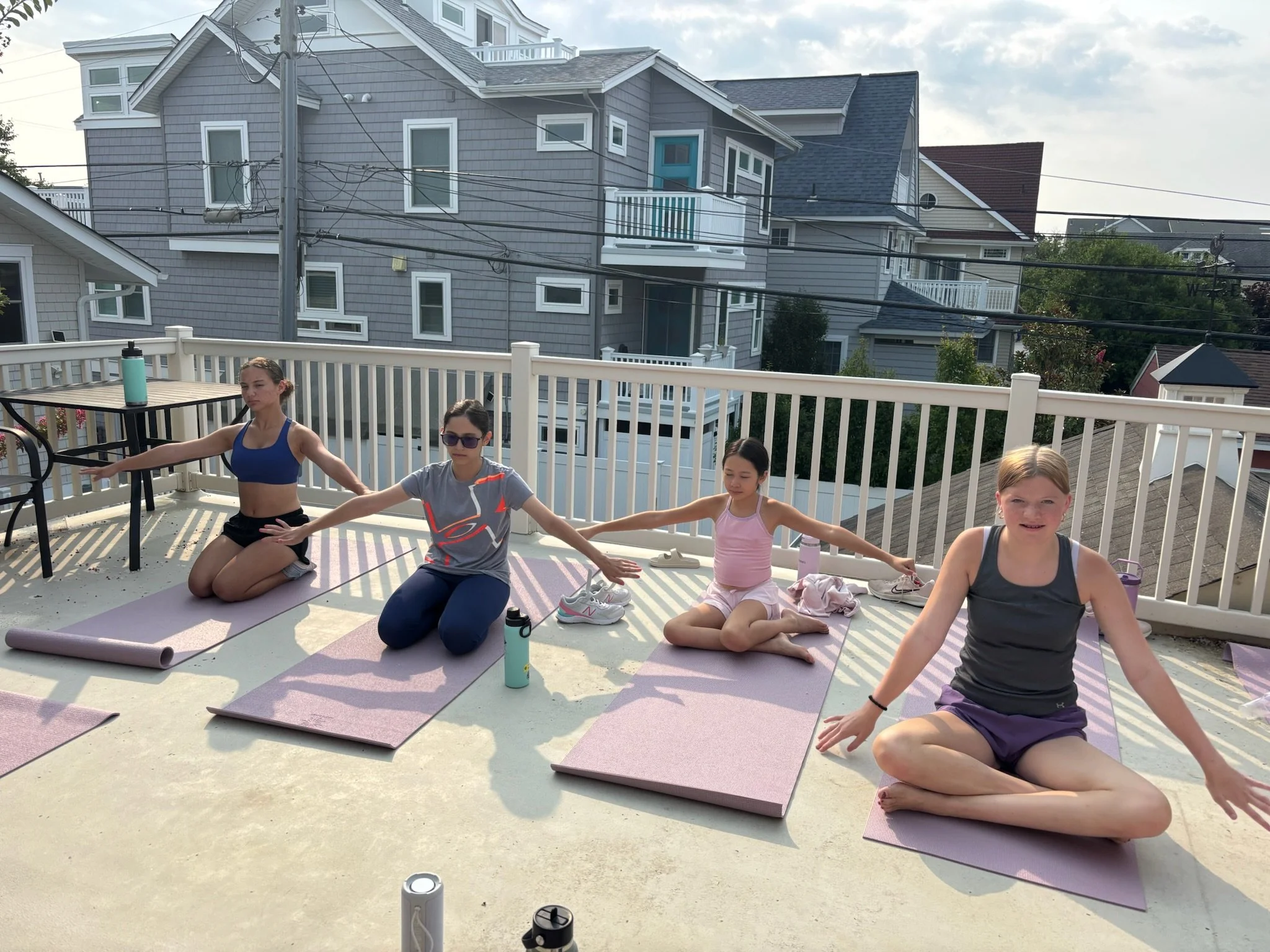 People doing yoga outdoors on a balcony with pink and purple yoga mats, surrounded by a white railing, with residential houses and a cloudy sky in the background.