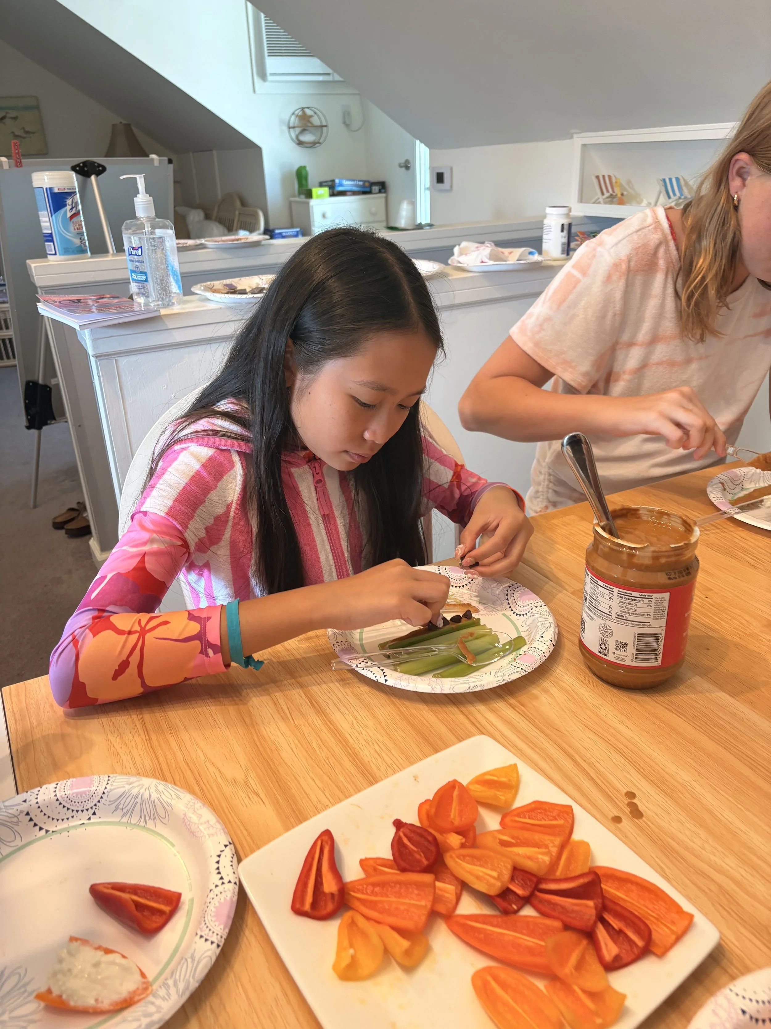 Two children at a dining table preparing or eating food, with a plate of sliced bell peppers in the foreground, and jars of condiments and plates on the table.
