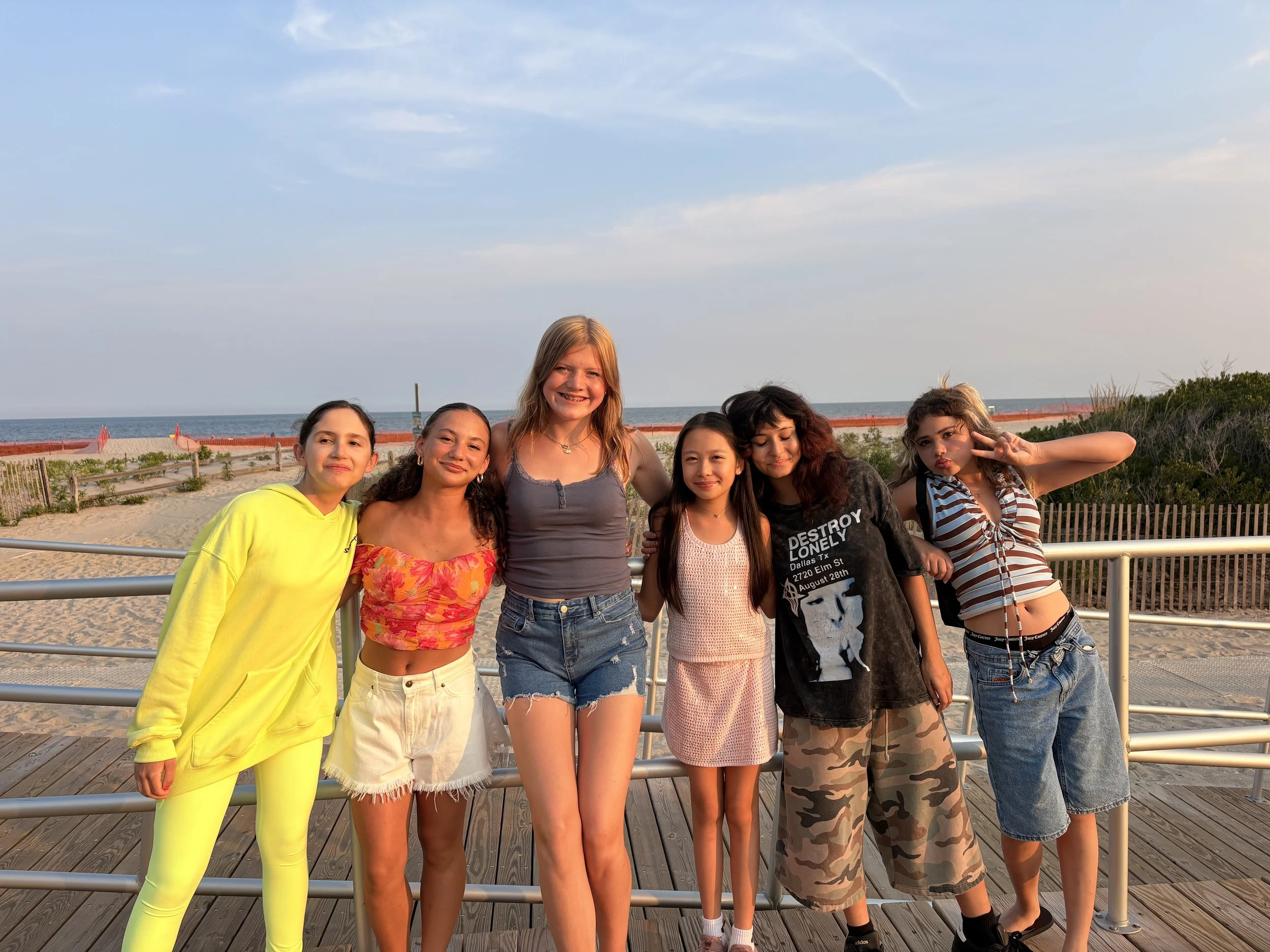 A group of six diverse young women posing together on a wooden walkway at a beach, with the ocean and a cloudy sky in the background.
