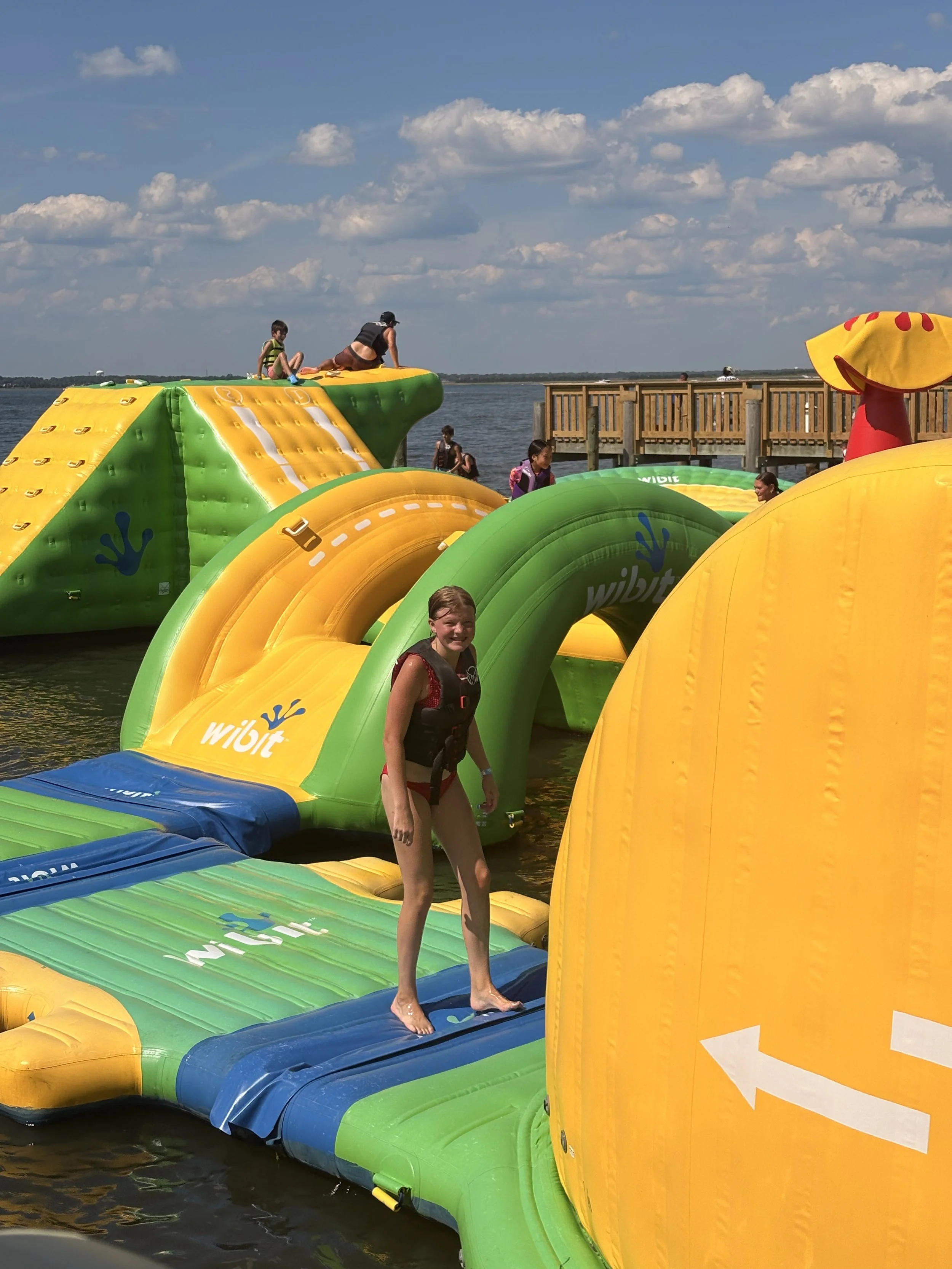 People enjoying themselves on a large inflatable water obstacle course on a lake, with a wooden pier and a partly cloudy sky in the background.