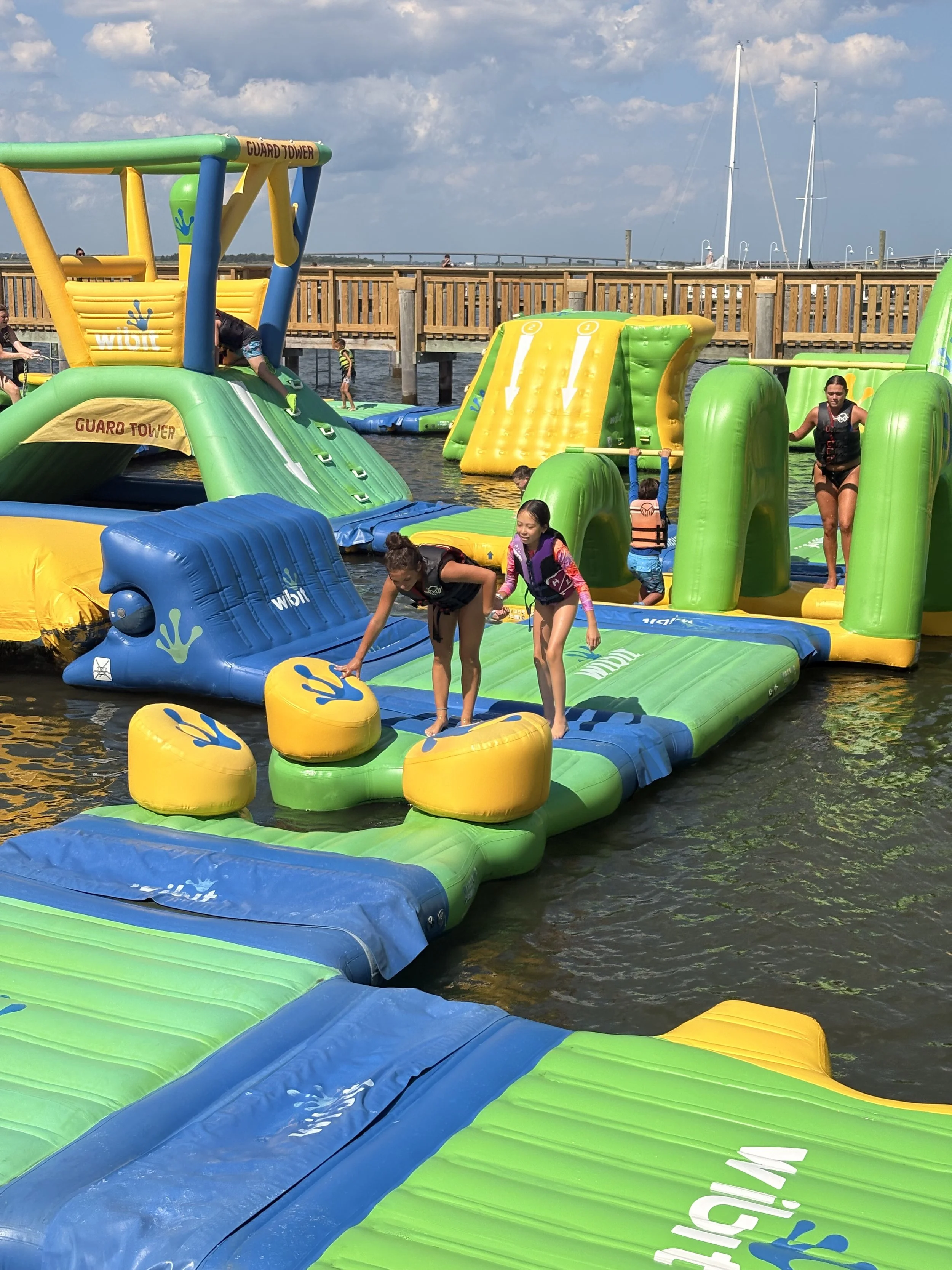 Children and adults enjoying themselves on inflated floating water obstacle course near a dock, with sailboats in the background and a partly cloudy sky.