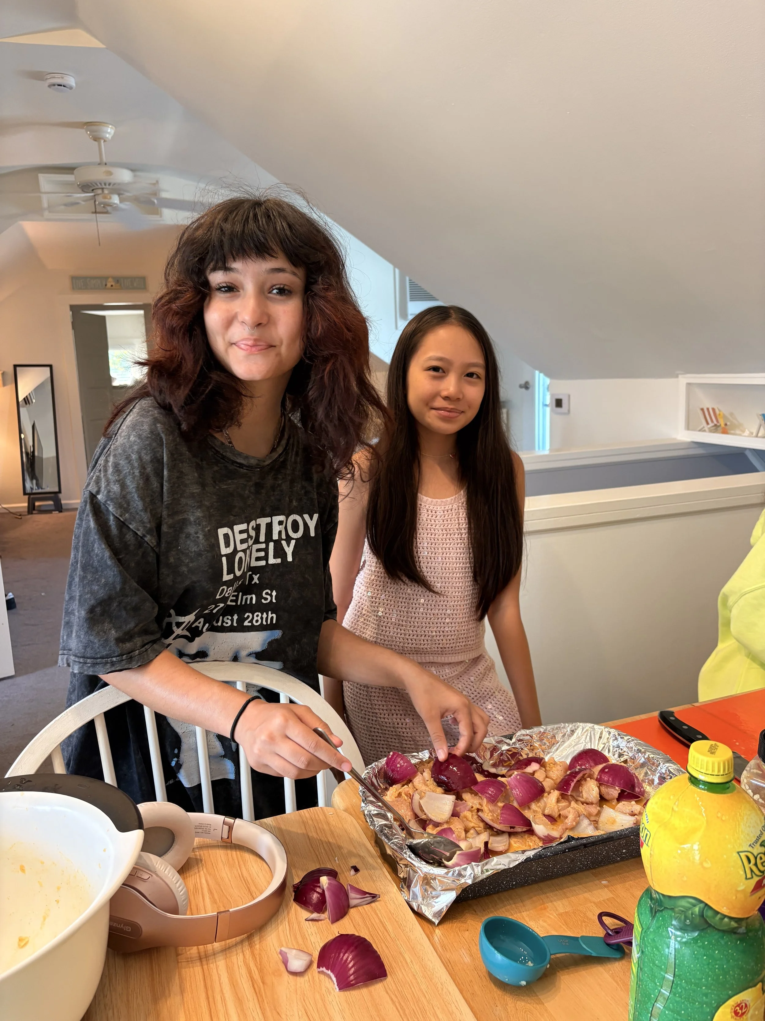 Two young women are preparing food in a kitchen. One is holding a utensil over a baking tray filled with chopped onions and beets. The table has various kitchen items, including a large mixing bowl, a pink headband, and a yellow bottle of lemon-lime 
