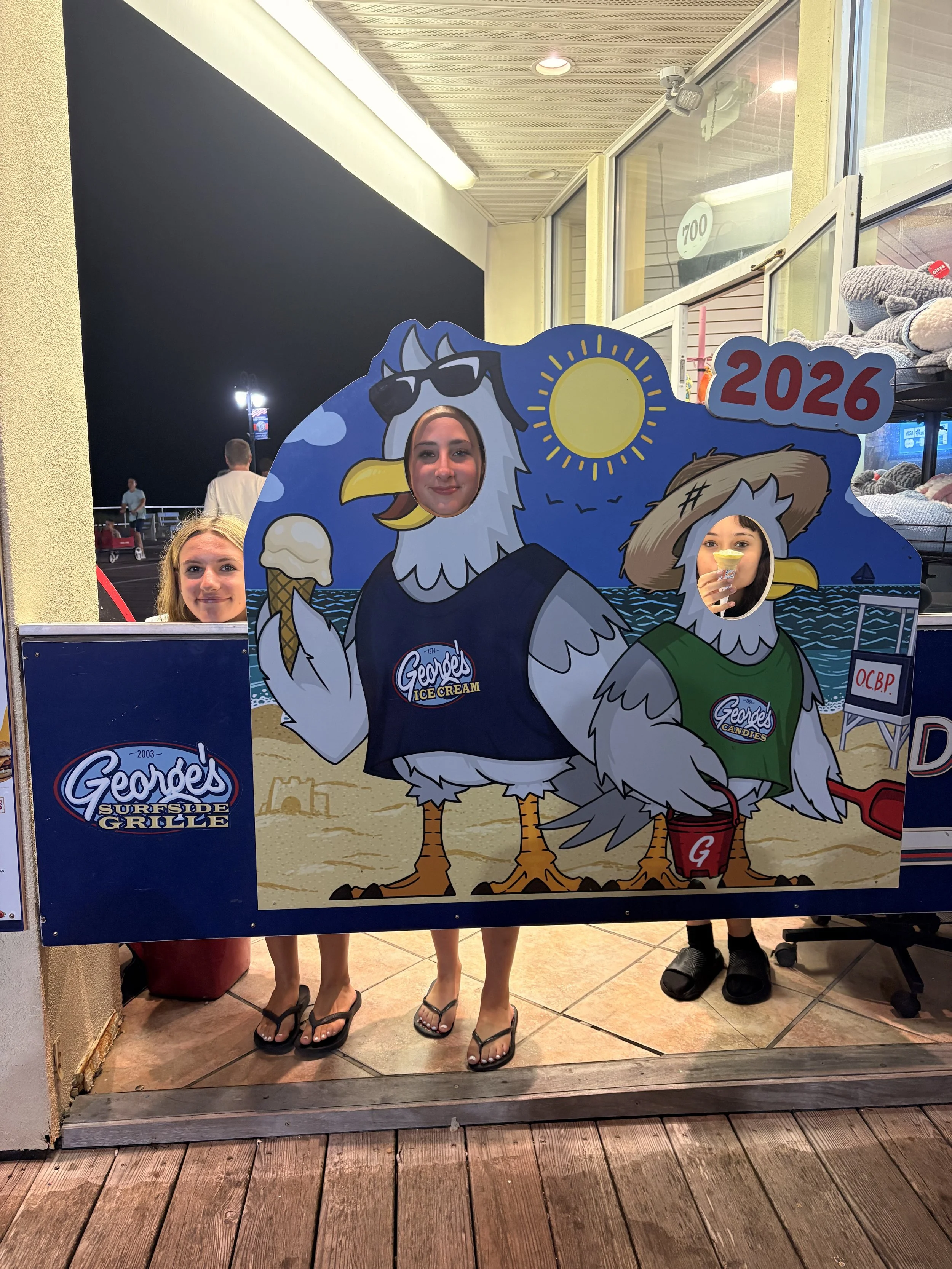 Three women in flip flops stand behind a Garde's Ice Cream cutout with cartoon illustrations of seagulls. The women are peeking through holes in the cutout, which depicts two seagulls wearing sunglasses, one holding an ice cream cone, and the other h