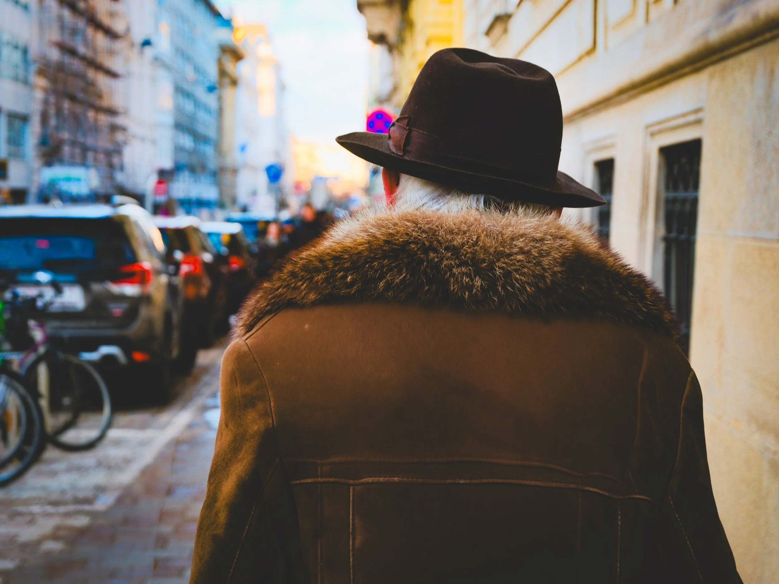 Person wearing a black hat and a coat with fur collar walking on city sidewalk with cars and bicycles parked along the street.