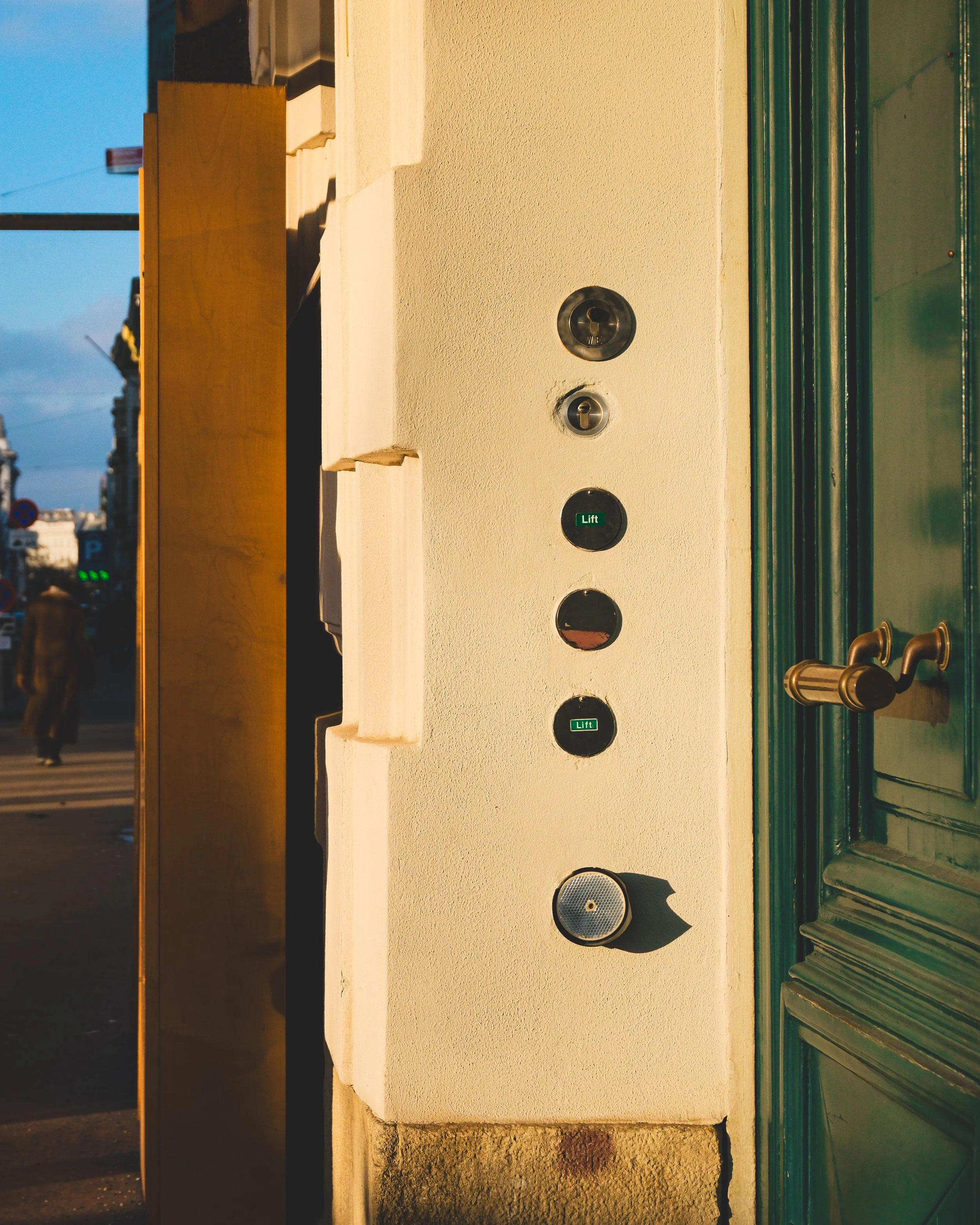Close-up of an intercom panel with multiple buttons and indicators, situated beside a green door with a brass handle, on a cream-colored wall outside a building during daytime.