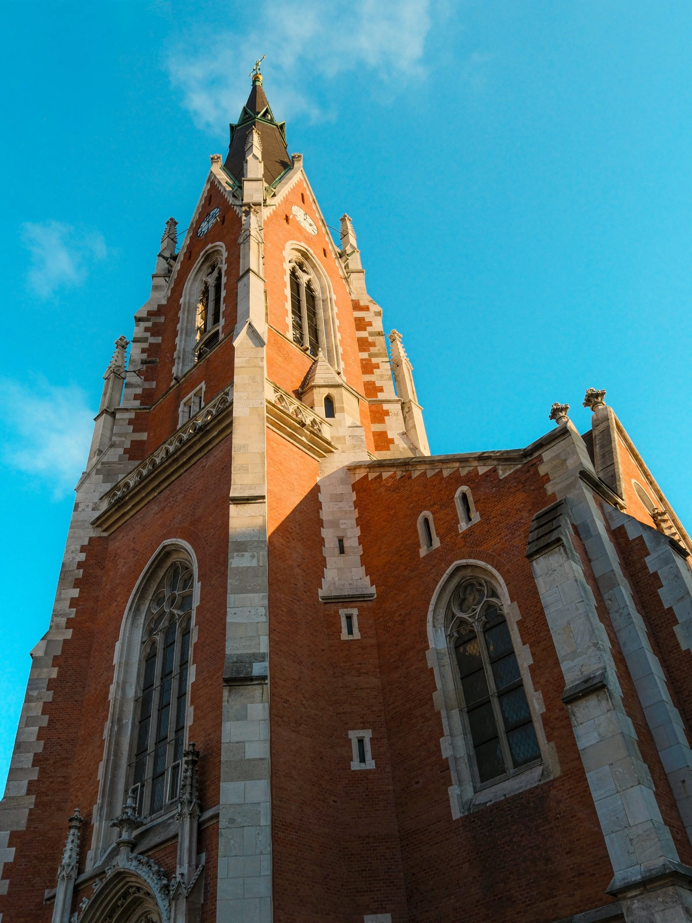 Low-angle view of a tall, ornate red brick church tower with Gothic architectural details and pointed arches against a blue sky.