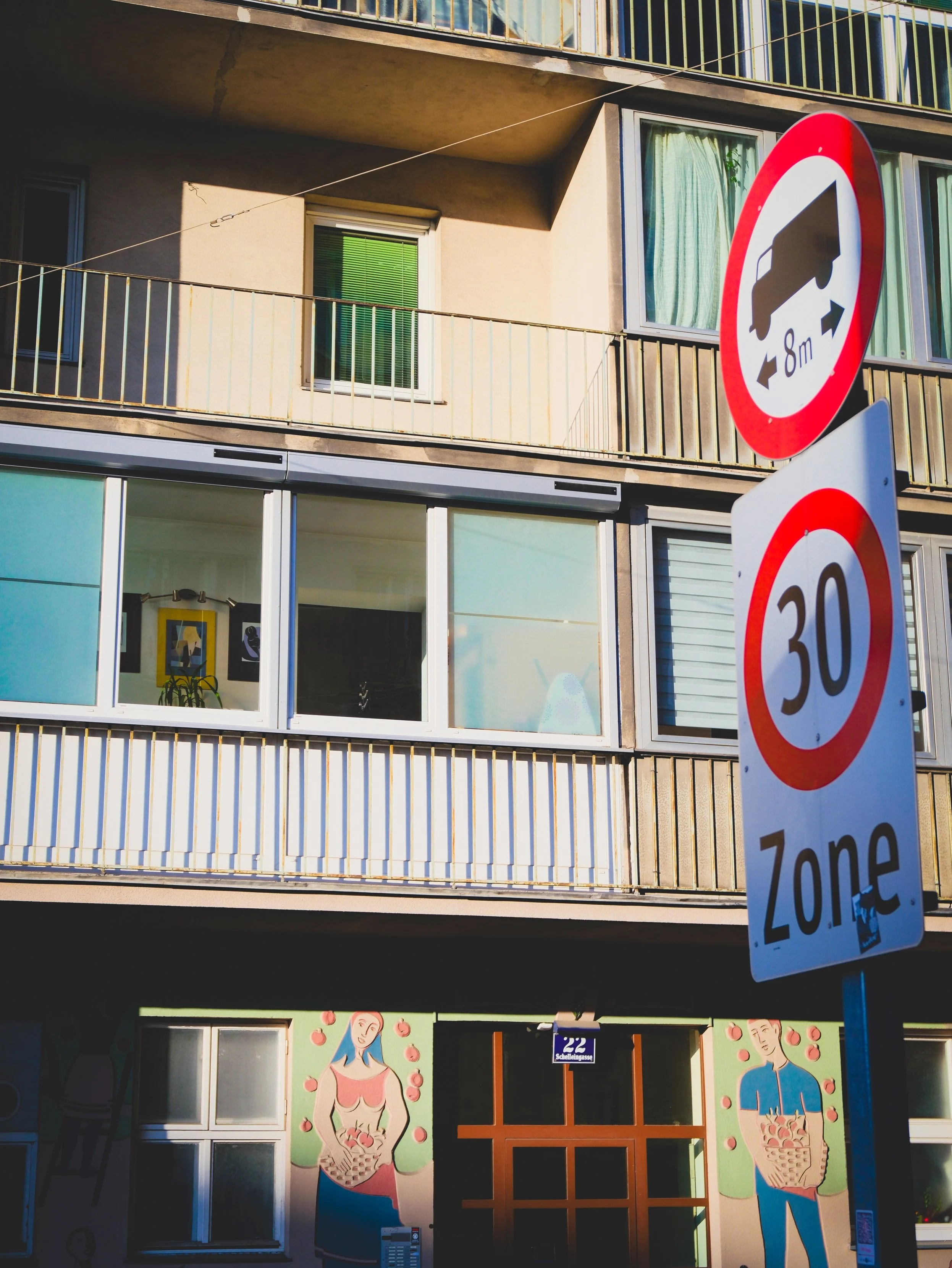 Multistory building with a mural of a woman and man holding baskets of fruit on the ground floor, and signs indicating an 8-meter distance restriction and 30 km/h speed limit zone in front.