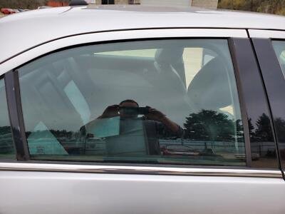 The side window of a silver car with a reflection of the sky and trees, and a person taking a photo.