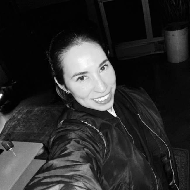 Black and white photo of a woman taking a selfie, smiling, with dark hair tied back, wearing a dark jacket, in an indoor setting with a carpeted floor and potted plant in the background.