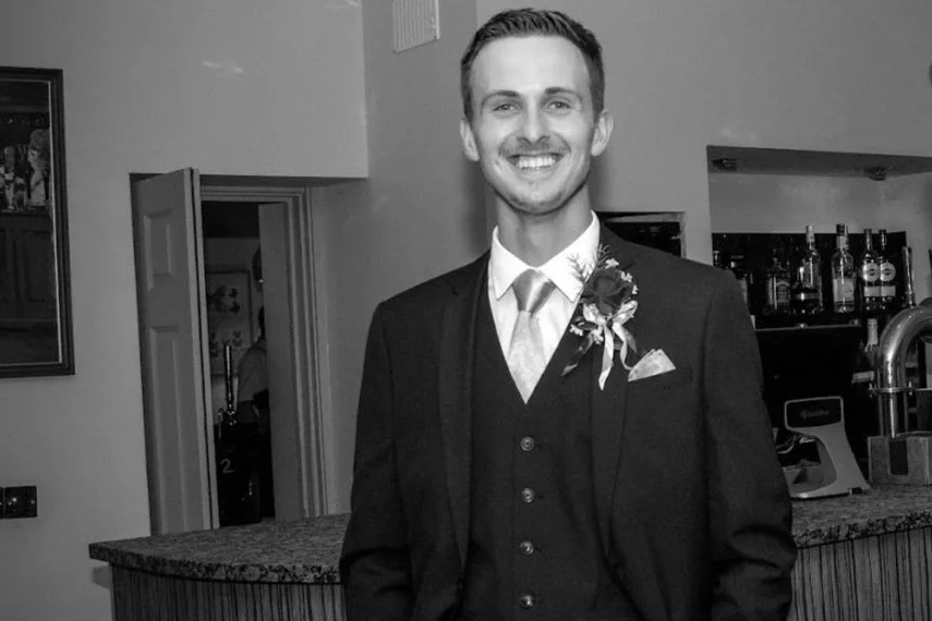 A young man with short brown hair wearing a dark jacket and black jeans stands on a balcony, overlooking the green space of a historic building with a city skyline in the background on an overcast day.