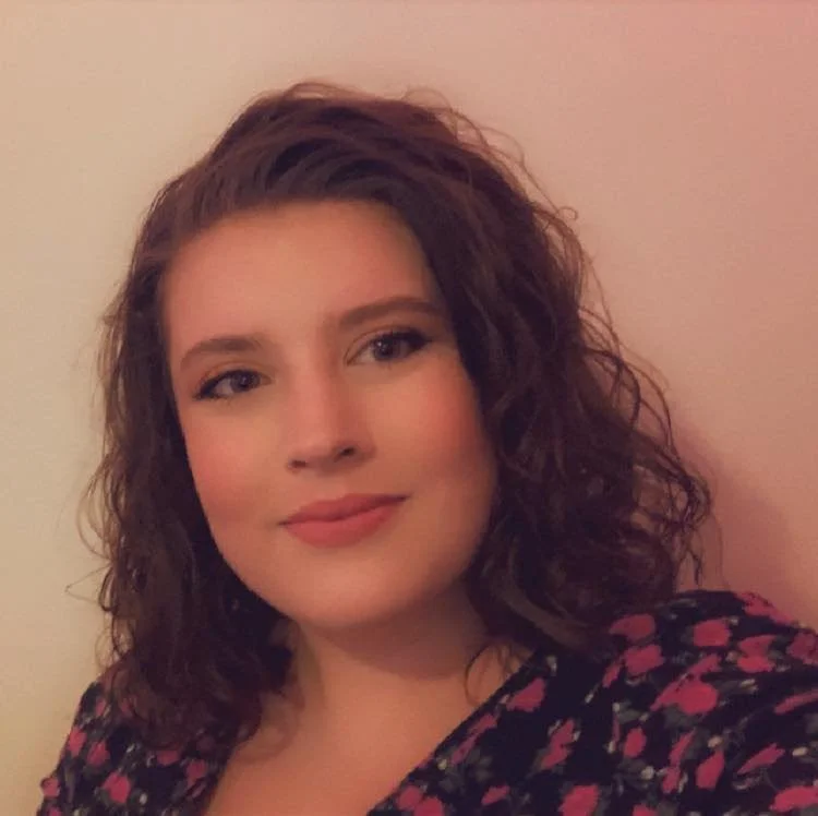 A young woman with shoulder-length curly brown hair, wearing a floral top, smiling in front of a light pink wall.
