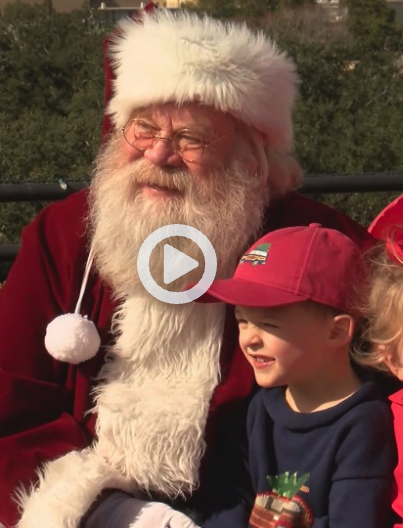 Santa Claus with a white beard and red suit sitting outdoors with children, one girl wearing a red hat and another child partially visible.